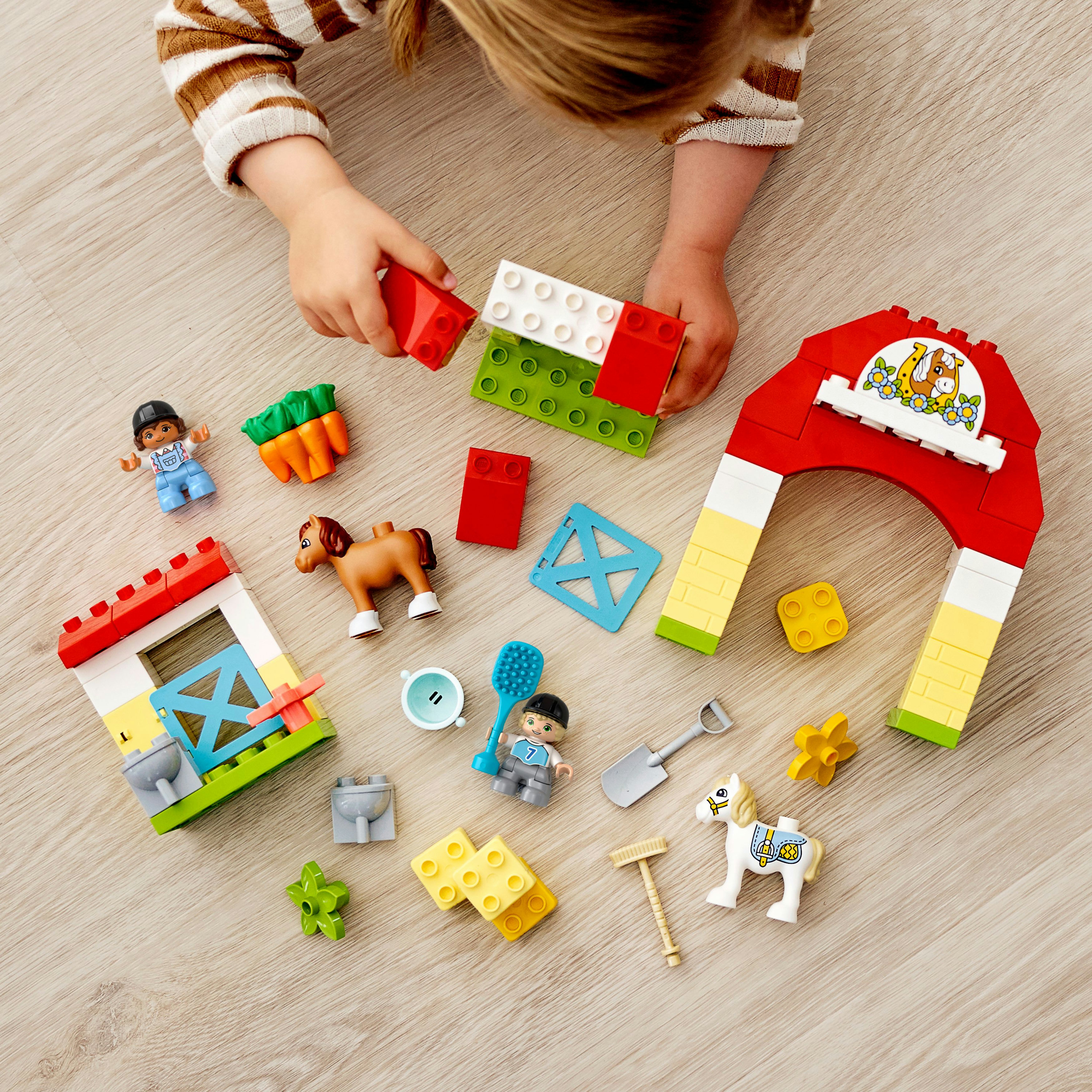 Bambino che gioca con il set fattoria Duplo; fienile rosso, figure e animali su pavimento in legno.