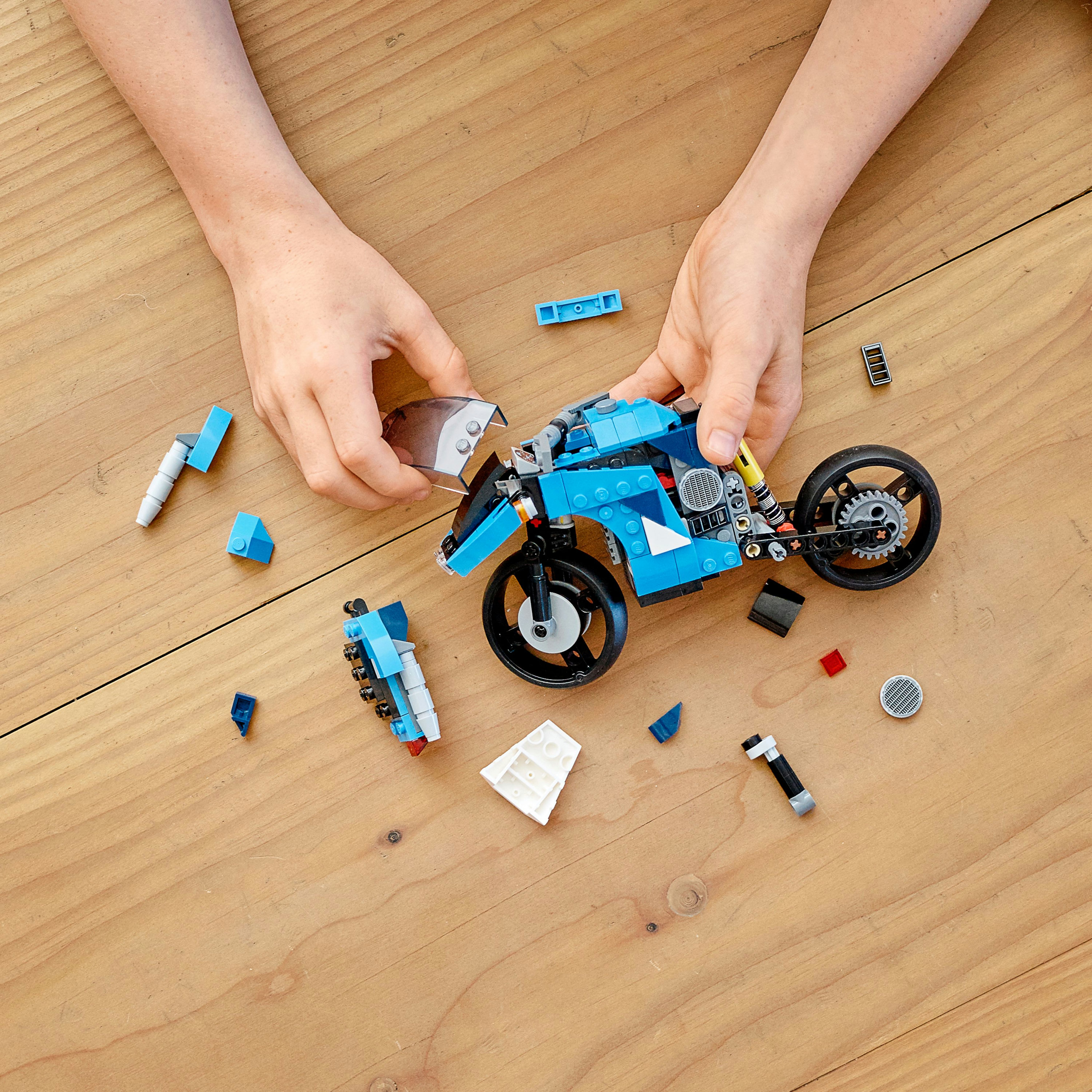 Hände, die ein blau-schwarzes Lego-Motorrad auf einer Holzoberfläche mit verstreuten Teilen zusammensetzen.