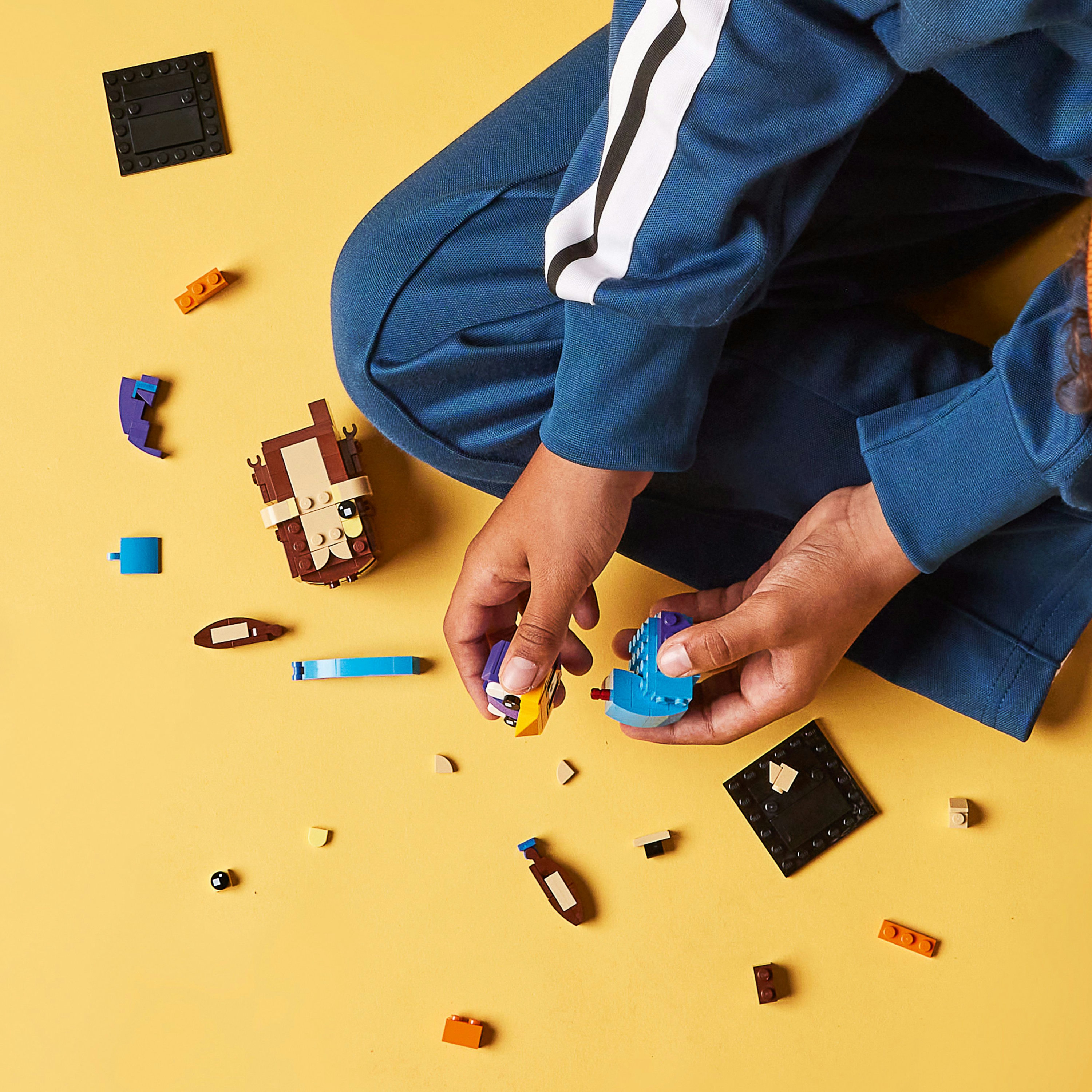 Bir çocuğun elleri sarı bir arka plan üzerinde iki LEGO yapımı tutuyor. Diğer LEGO parçaları dağınık.