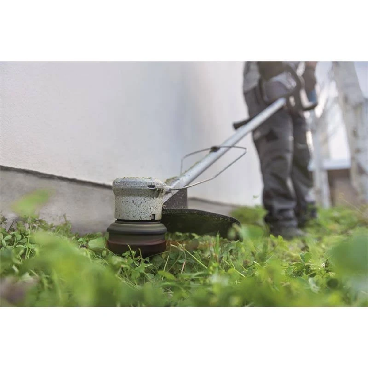 Una persona usa un cortabordes para cortar hierba cerca de una pared blanca, con hierba verde en primer plano.