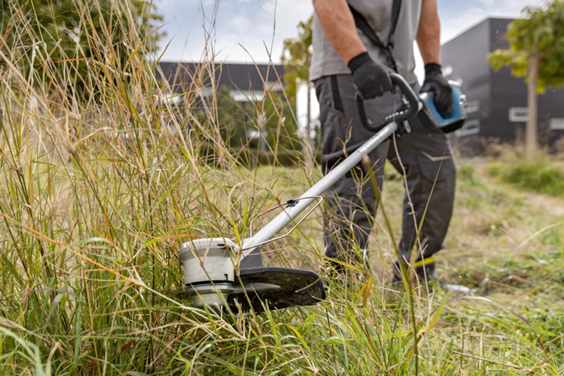 Ein Mann benutzt einen Fadentrimmer, um hohes Gras in einem grasbewachsenen Bereich zu schneiden. Der Himmel ist bedeckt.