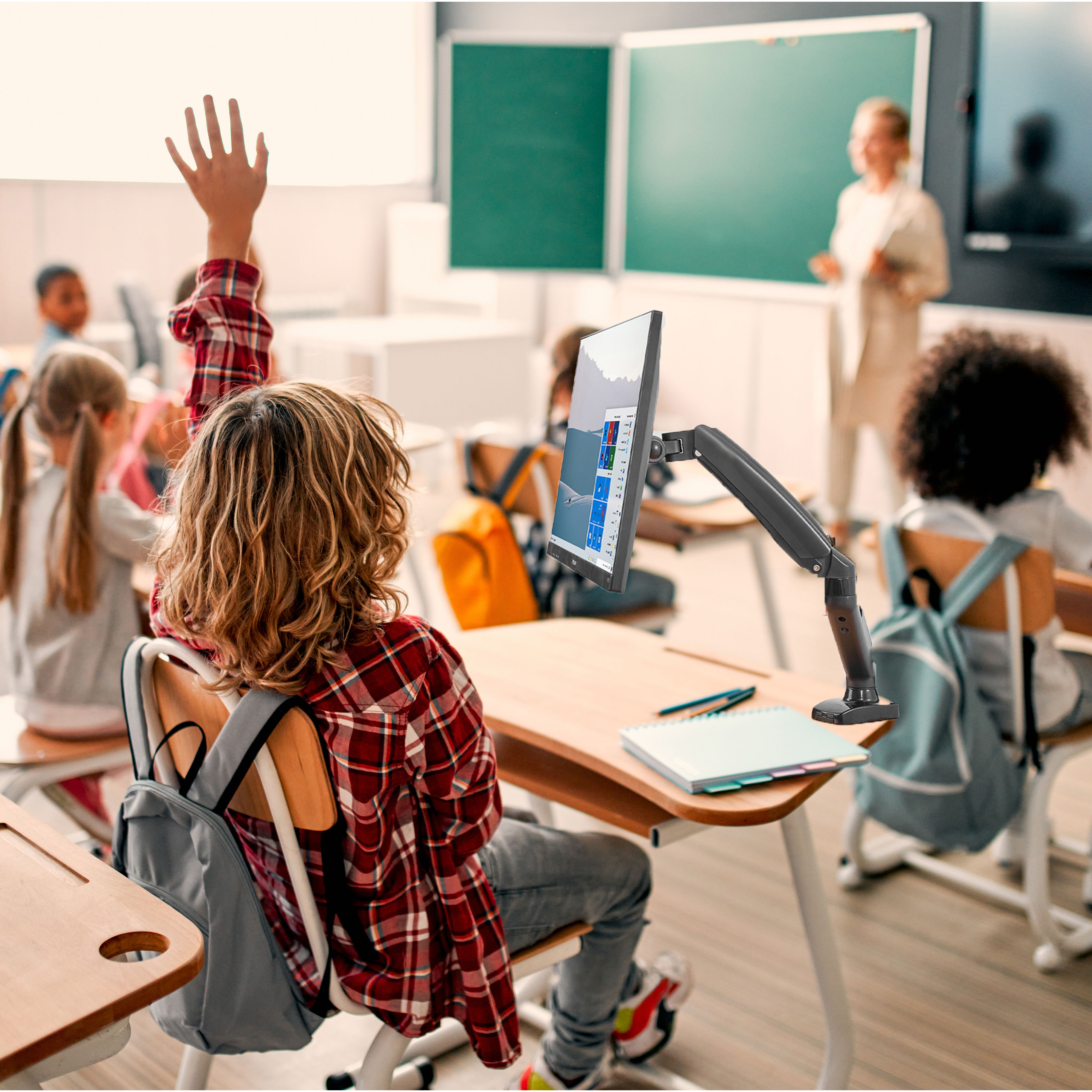 Ein Schüler hebt die Hand im Klassenzimmer. Lehrer und Schüler im Hintergrund. Monitor auf einem Schreibtisch.