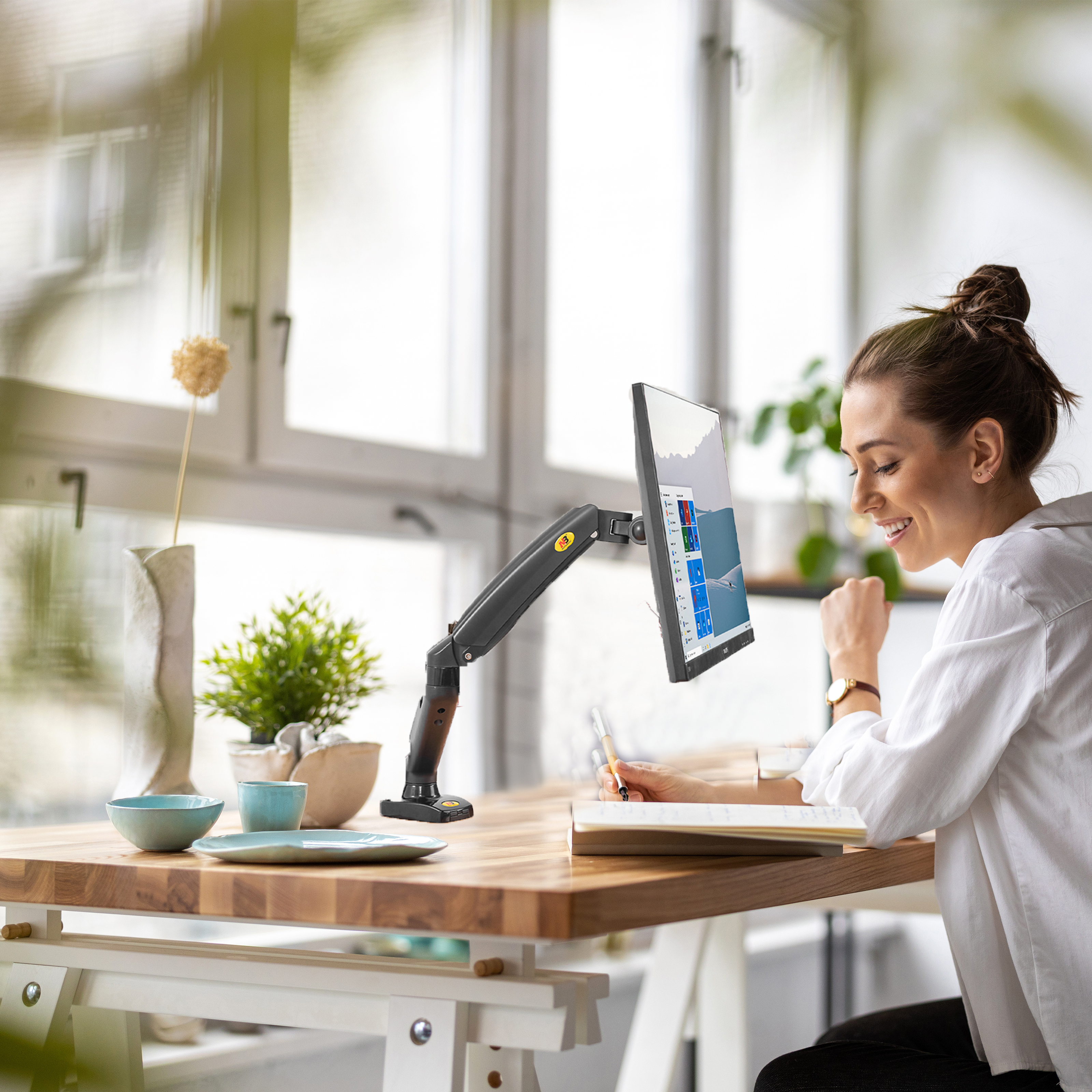 Eine Frau arbeitet an einem Schreibtisch mit einem Computermonitor an einem verstellbaren Arm. Sie lächelt und schreibt.