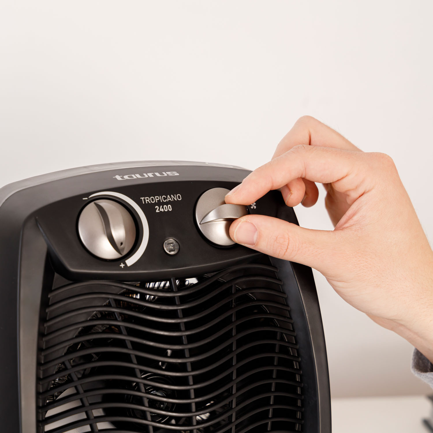 Ręka reguluje pokrętło wentylatora na czarnym grzejniku Taurus Tropicano, na białym tle.