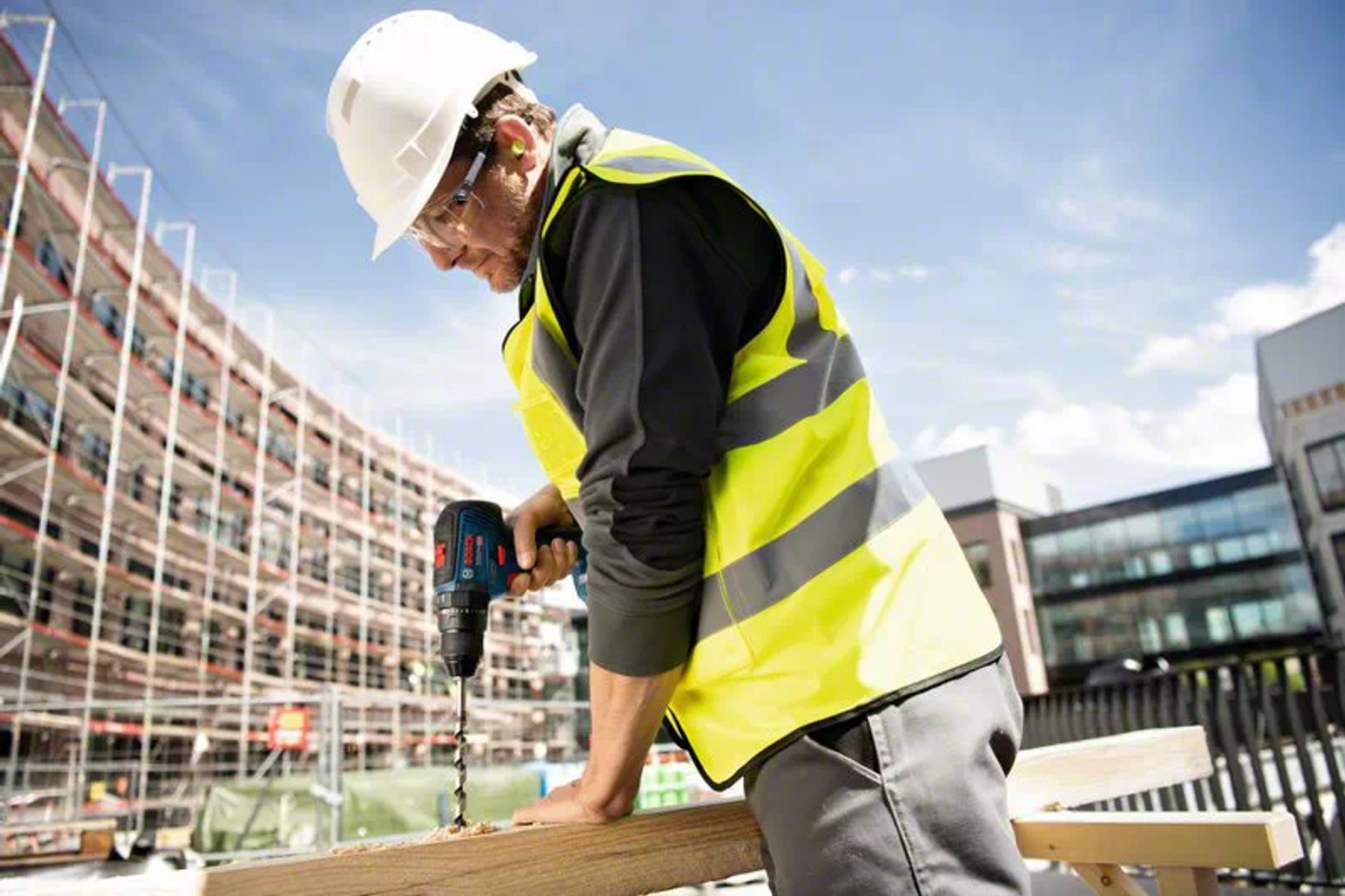 Mężczyzna w kamizelce ochronnej używa wiertarki na drewnianej desce na zewnątrz, pod błękitnym niebem.
