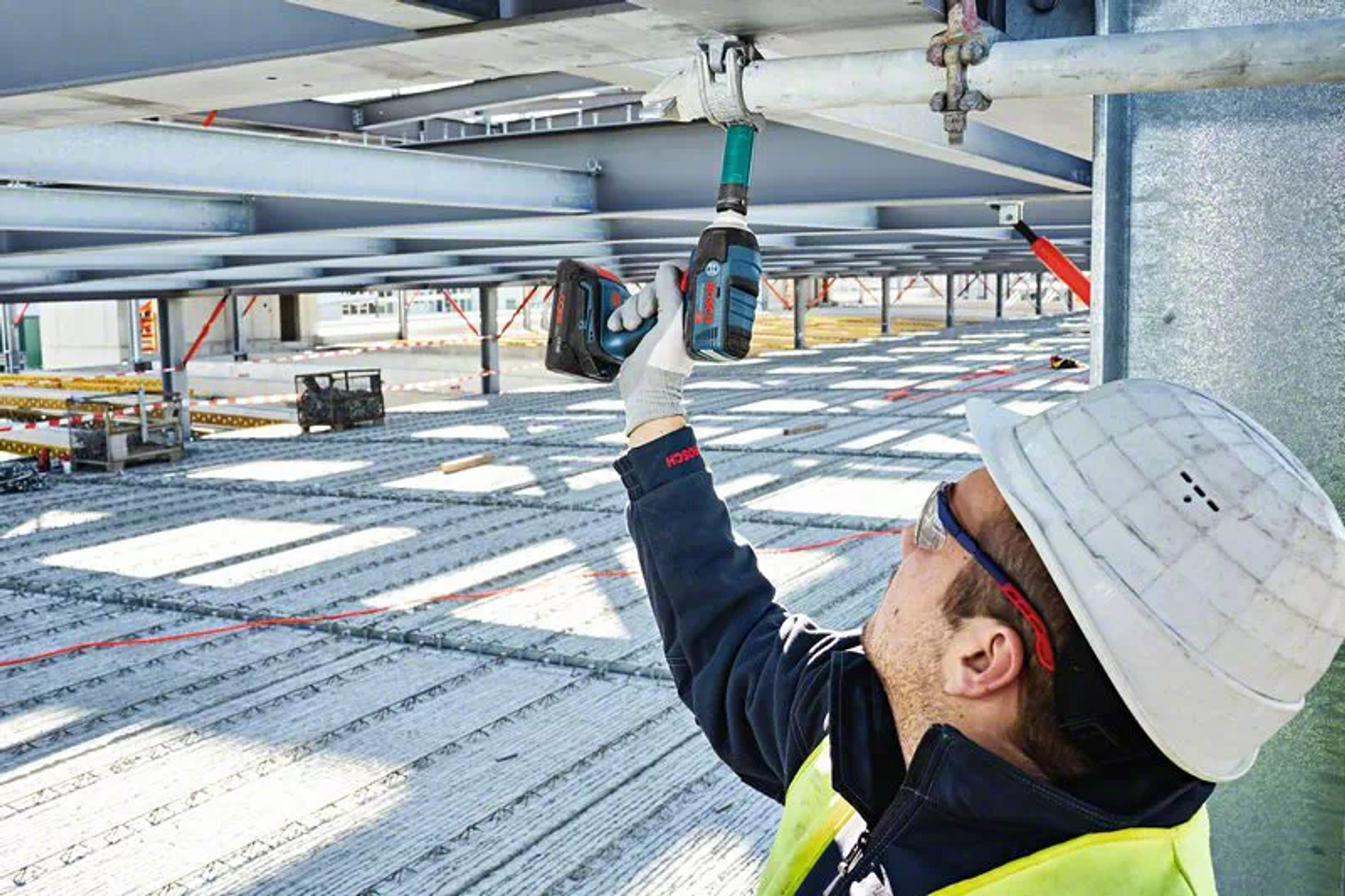 Mężczyzna w kasku używa elektronarzędzia do mocowania metalowej belki na budowie.
