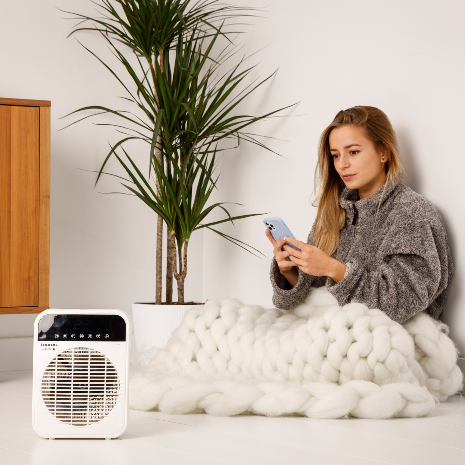 Kobieta siedzi na białym kocu, używając telefonu w pobliżu grzejnika i rośliny.