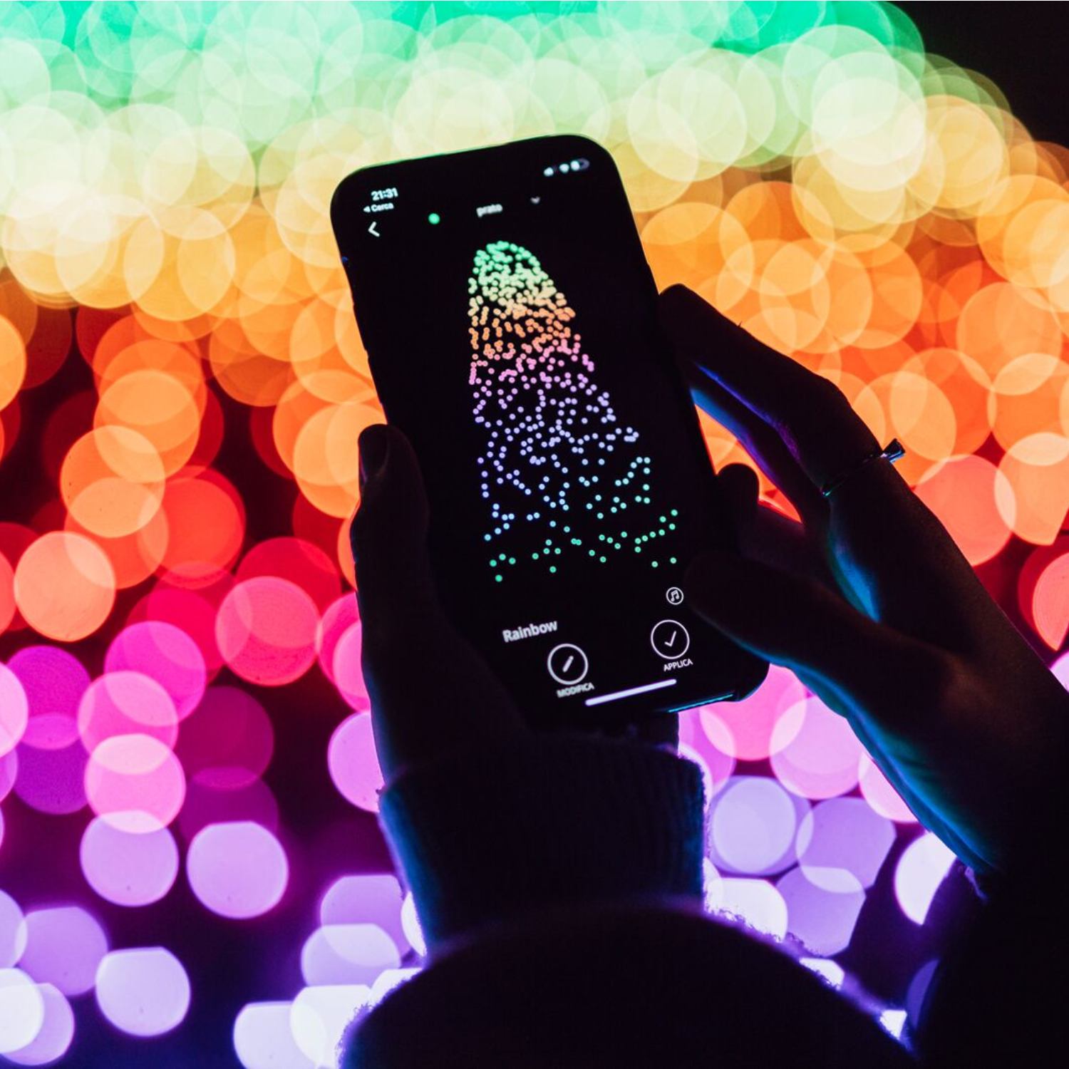 Telefon wyświetla kolorowy wzór tęczy. Rozmyte światła w tle. Ręce trzymające telefon.