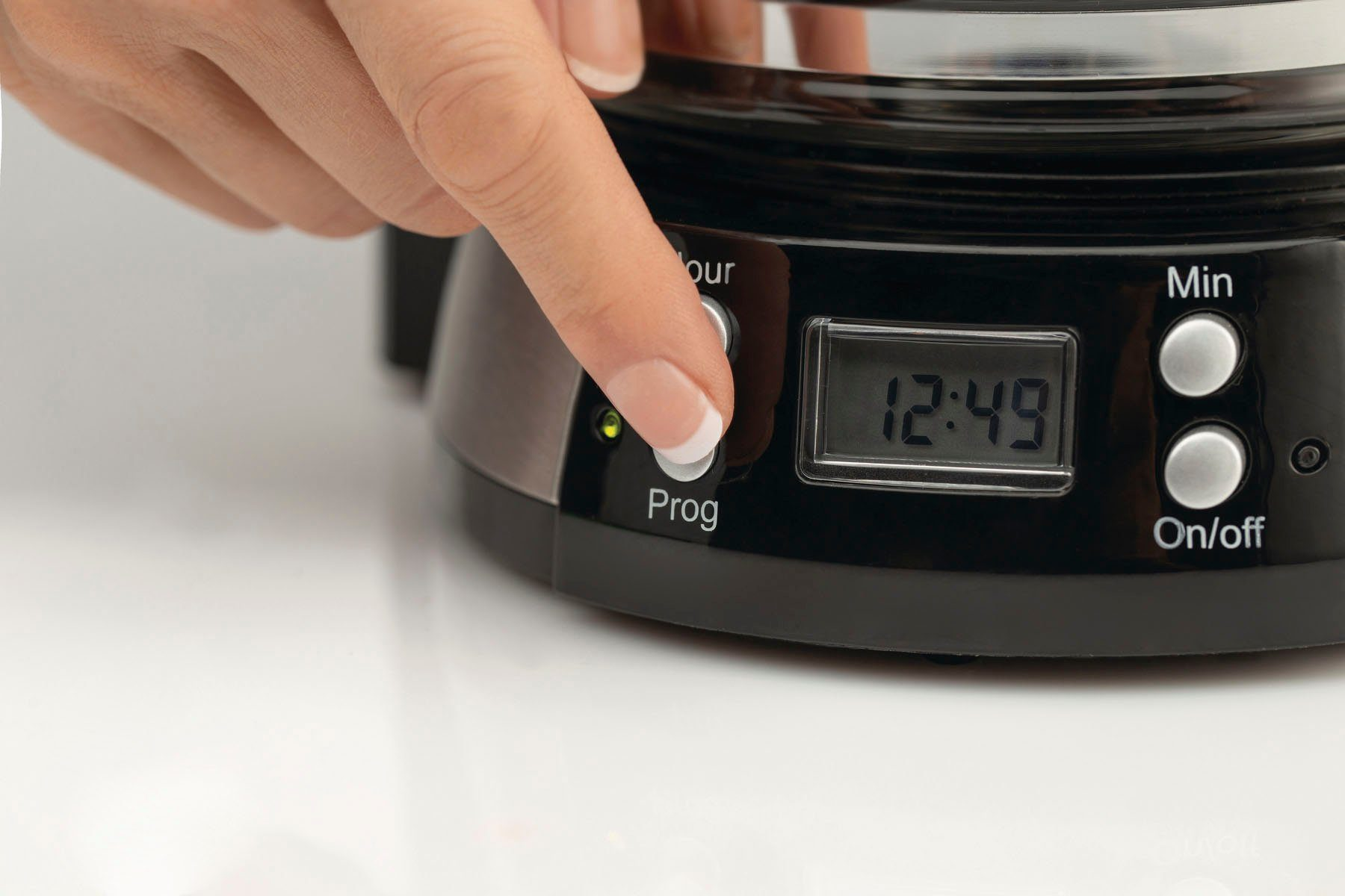 Nahaufnahme einer Hand, die eine Taste an einer Kaffeemaschine drückt. Die Anzeige zeigt 12:49. Andere Tasten sagen 'Stunde', 'Min'.