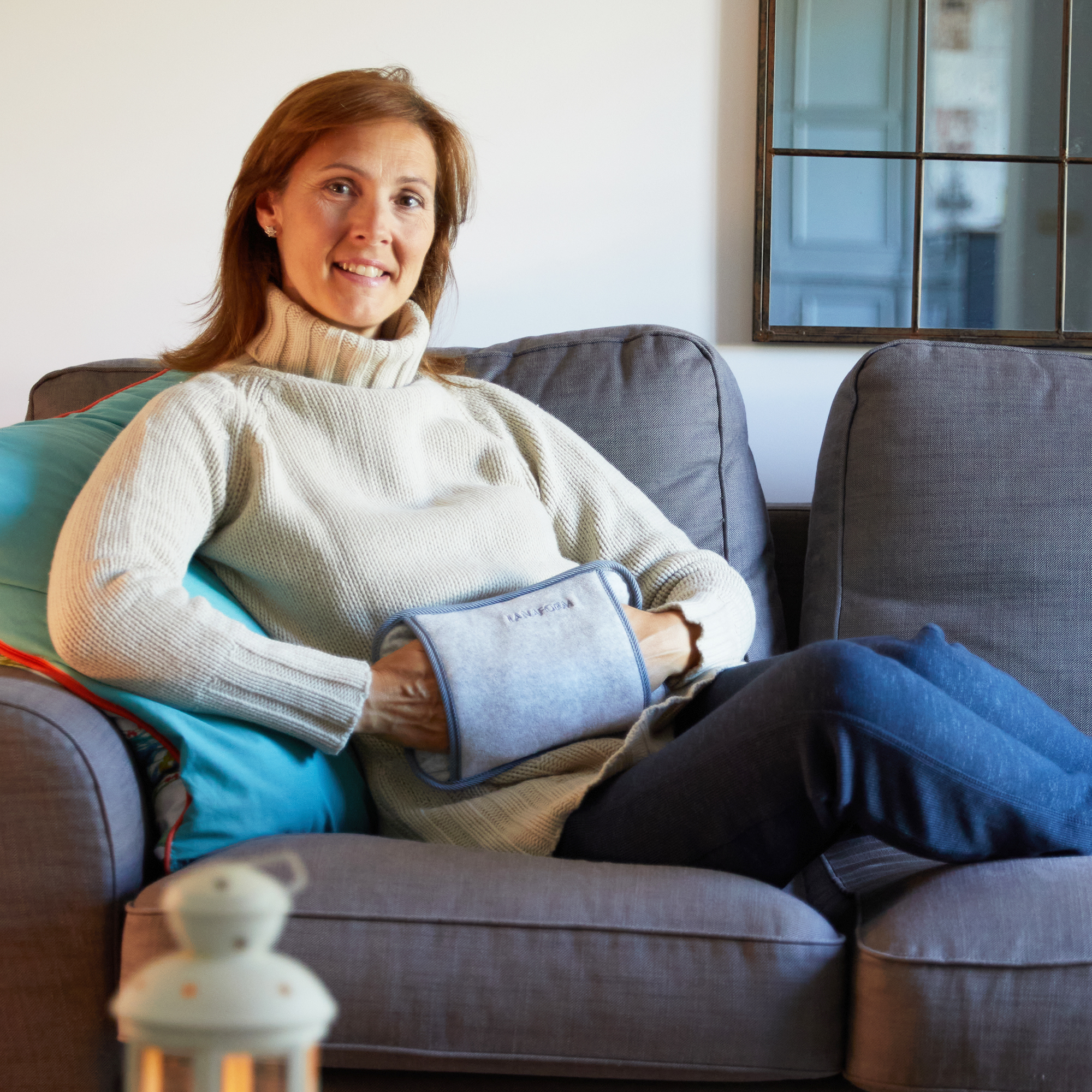 Frau auf grauem Sofa hält graue Wärmflasche, lächelt, mit blauem Kissen.