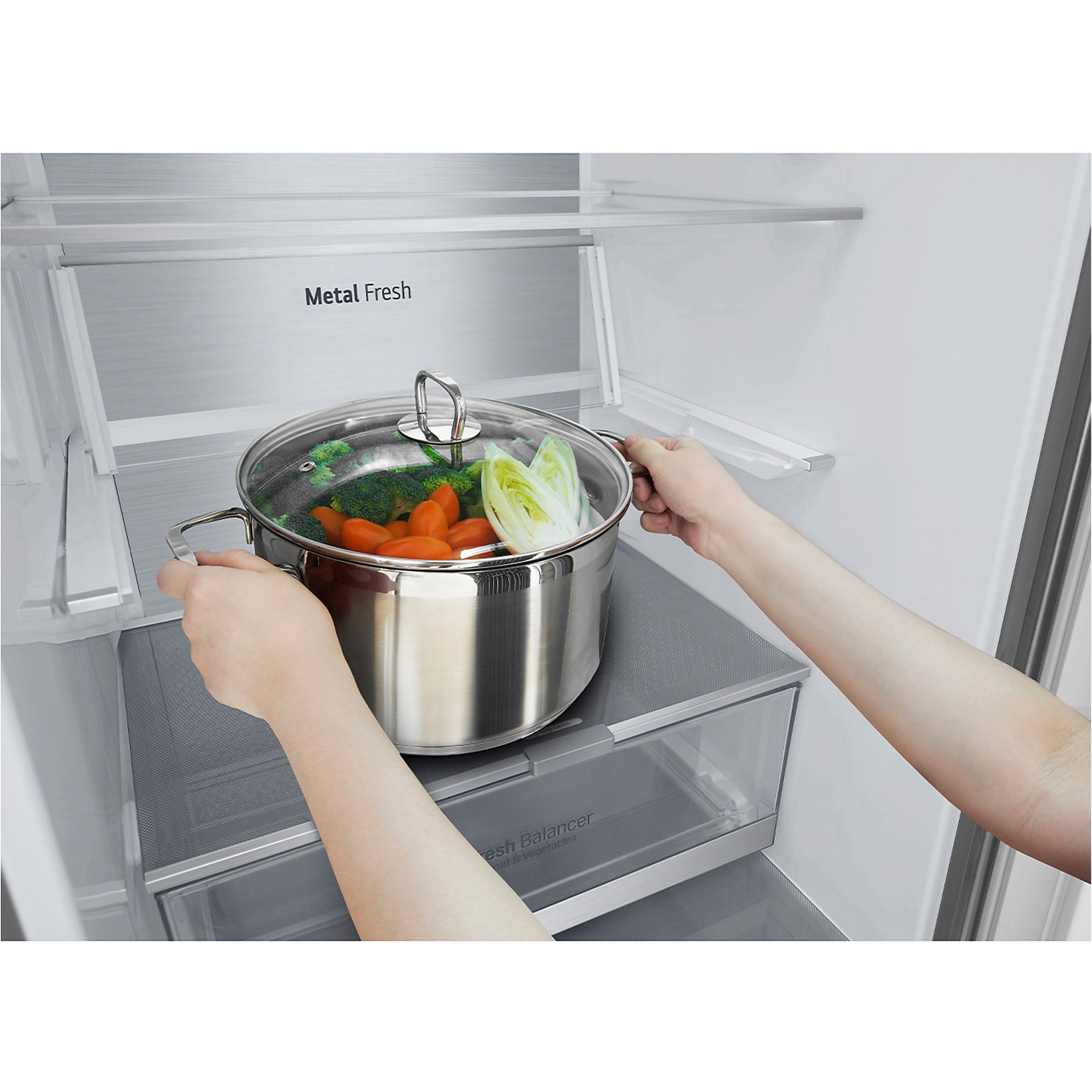 Dos manos colocan una olla con verduras dentro de un refrigerador. Se ven estantes de metal.
