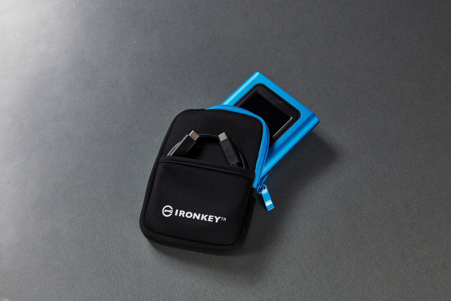 Czarny futerał Ironkey z zamkiem błyskawicznym. Niebieskie urządzenie częściowo widoczne. Na szarej powierzchni.