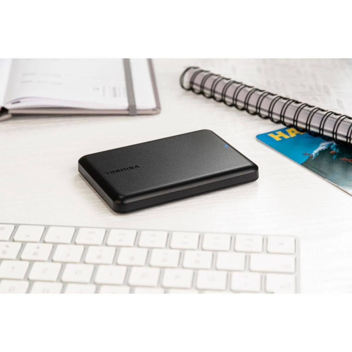 Un disco duro externo Toshiba negro en un escritorio, con teclado, cuaderno y una tarjeta.