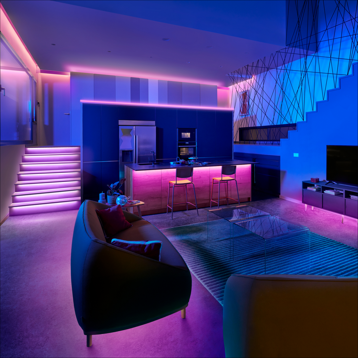 Modern living room with blue and pink LED lighting. Couch, stairs, kitchen, and cabinets with lights.