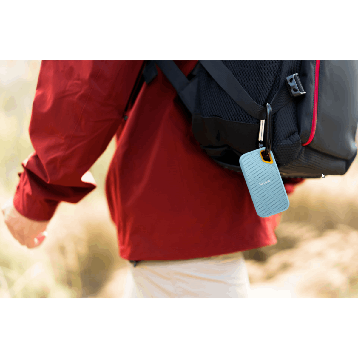 Osoba w czerwonej kurtce idąca z jasnoniebieskim zewnętrznym dyskiem twardym na plecaku.