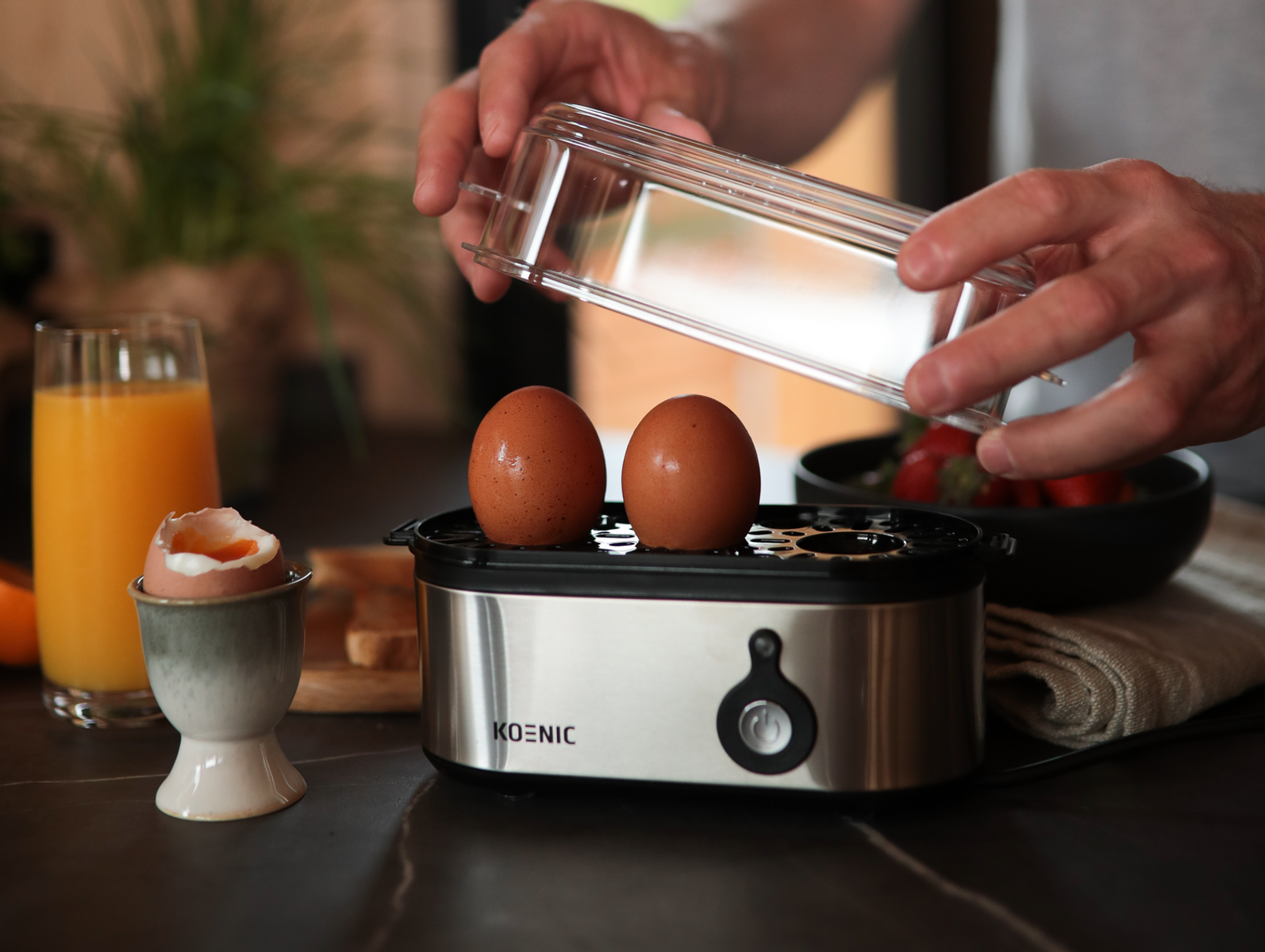 Eine Person öffnet einen durchsichtigen Deckel über zwei braunen Eiern in einem Eierkocher. Orangensaft daneben.