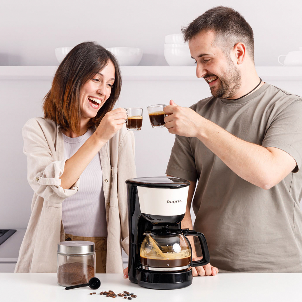 Un hombre y una mujer brindan con tazas de café. Una cafetera está en el mostrador y granos de café debajo.