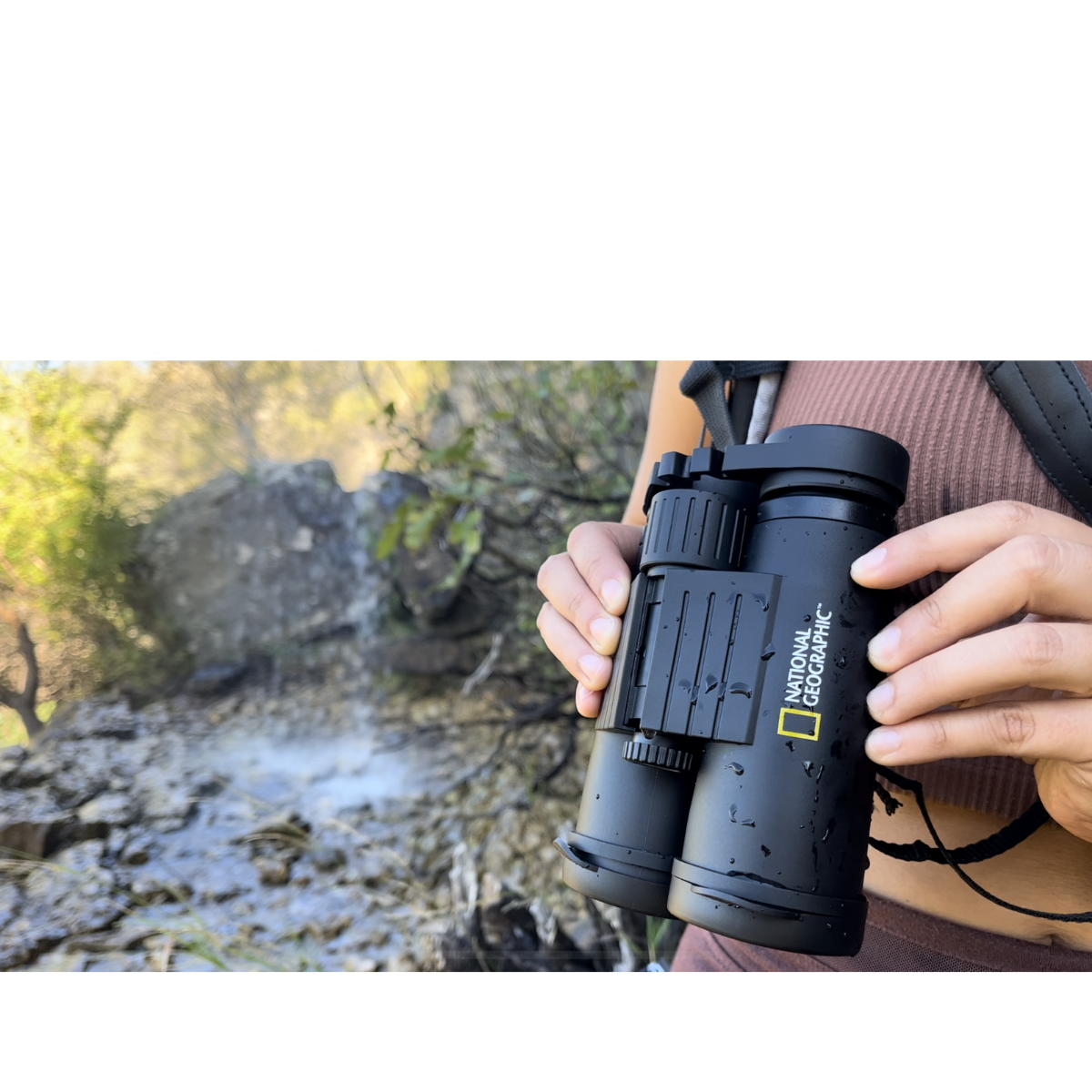 Persona sosteniendo binoculares negros cerca de una cascada. Los binoculares tienen una correa y un logotipo.