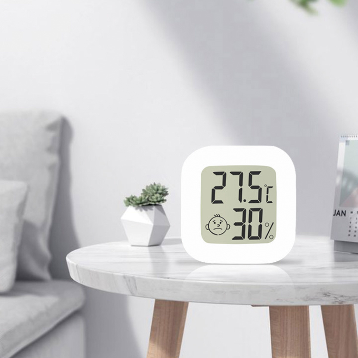 Ein weißes digitales Thermometer zeigt 27,5°C und 30 % Luftfeuchtigkeit auf einem Tisch mit Pflanze.
