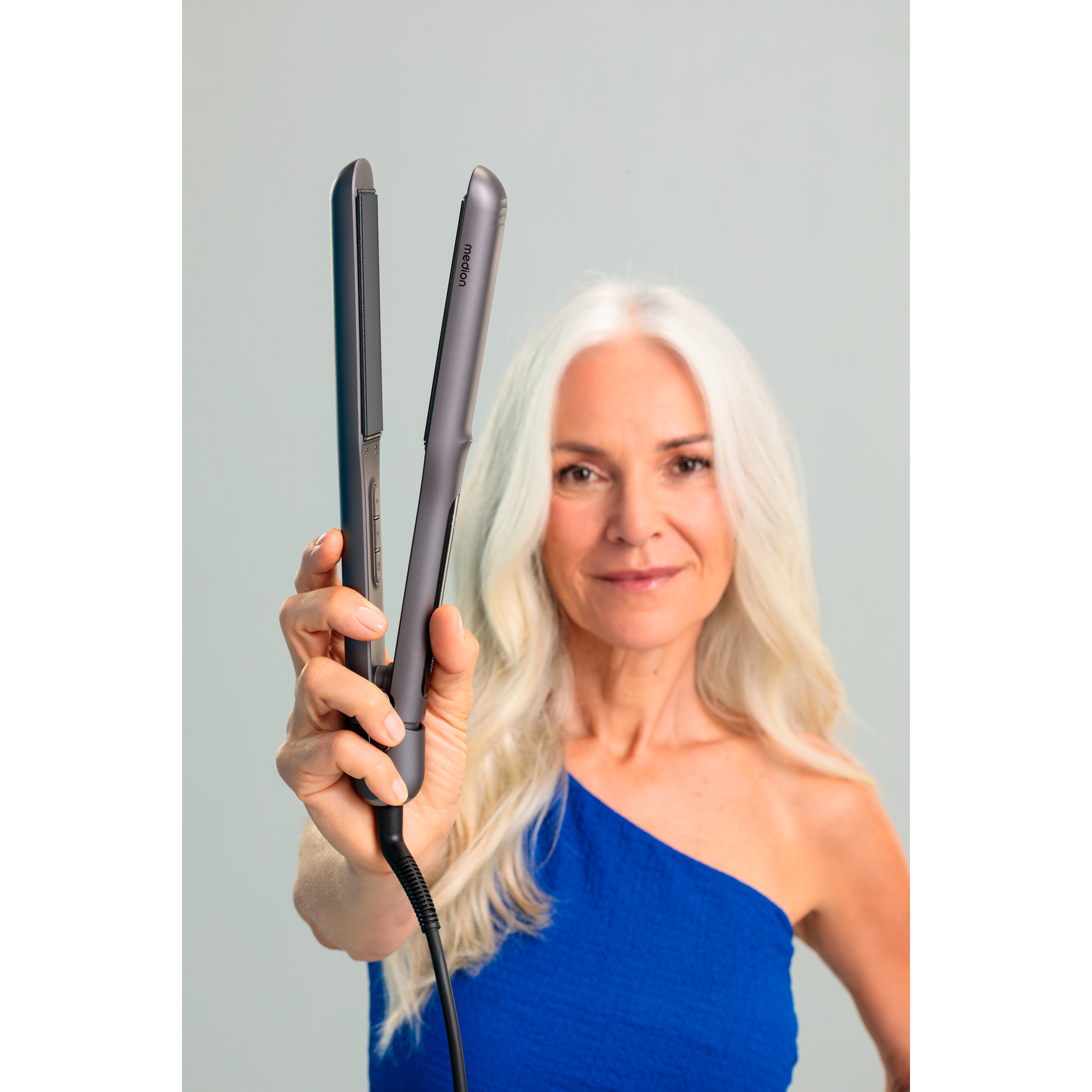 Kobieta z długimi, białymi włosami, trzyma prostownicę. Ma na sobie niebieską sukienkę i uśmiecha się. Jasne tło.