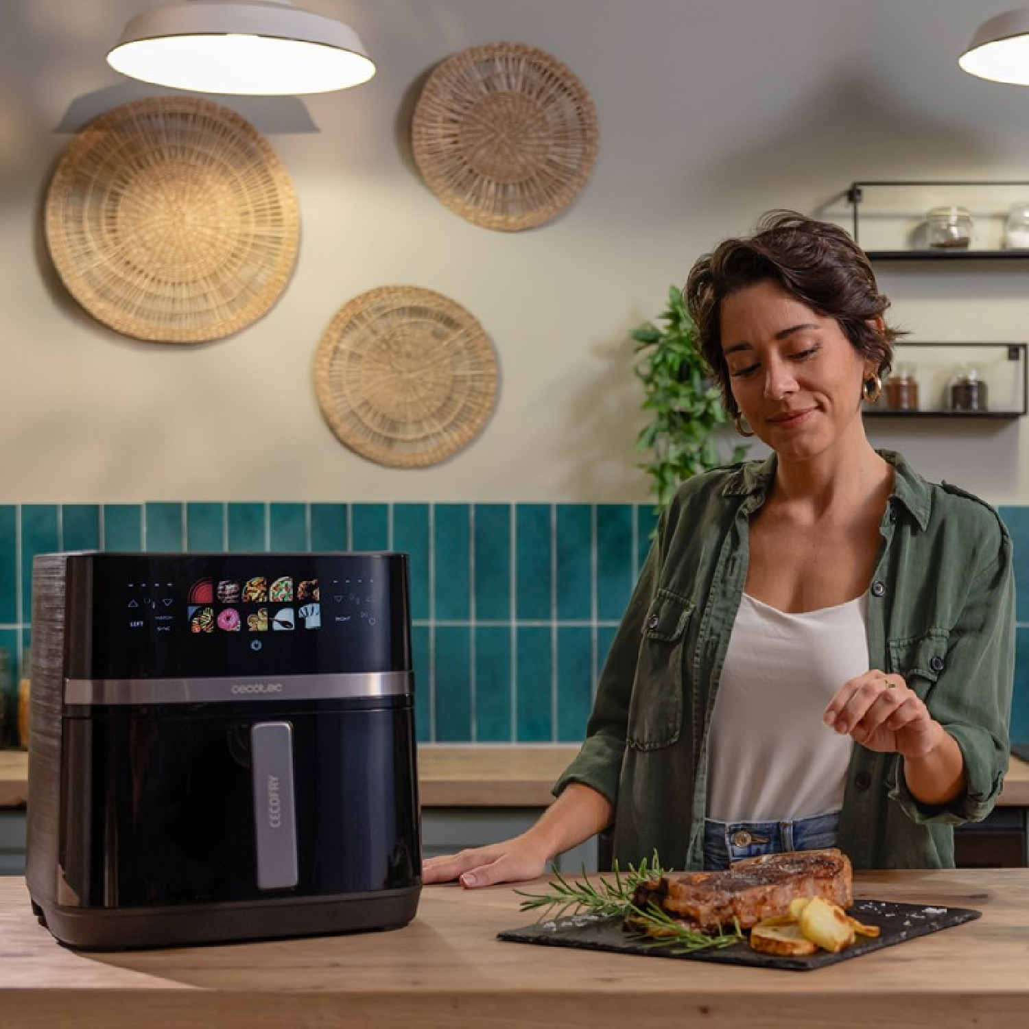 Kobieta w kuchni, w pobliżu frytkownicy beztłuszczowej. Stek na kamiennej desce. Ozdoby z wikliny na ścianie.