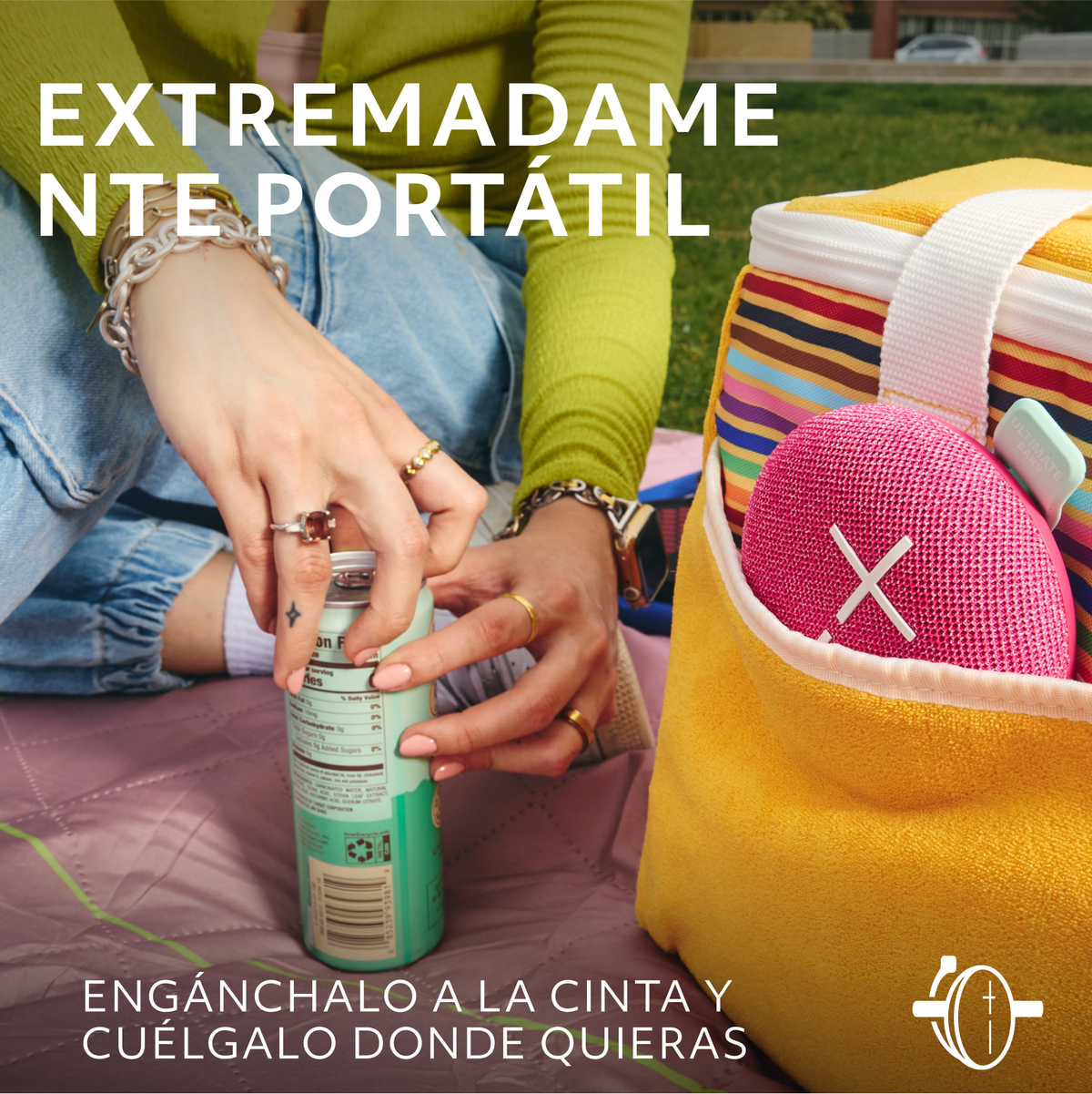 Osoba trzymająca napój, różowy głośnik w żółtej torbie, na kocu piknikowym.