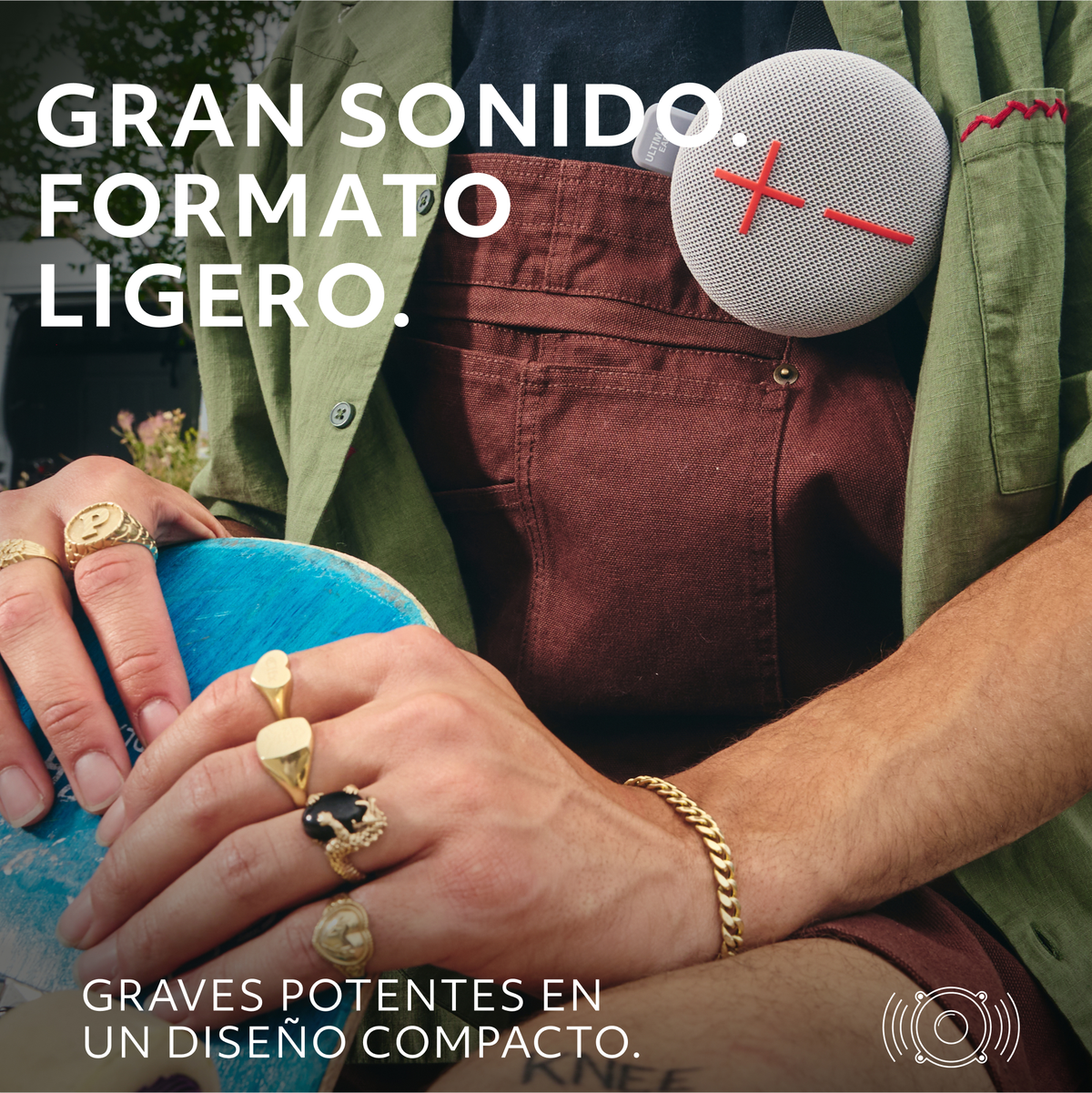 Ręce osoby trzymają mały, okrągły, szary głośnik. Mają na sobie pierścionki i złotą bransoletkę.