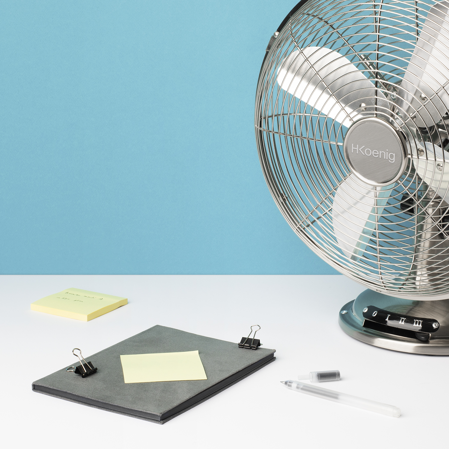 Un ventilatore HKoenig argentato su un tavolo bianco con post-it, fermagli e una penna su sfondo blu.