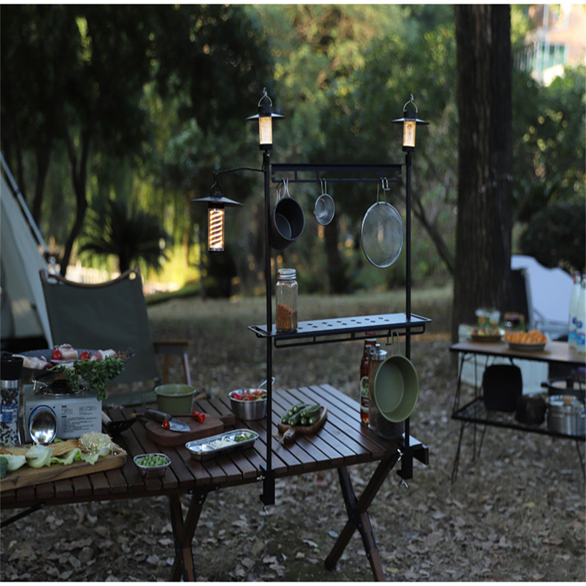 Camping-Setup mit Tisch, Essen und Kochausrüstung. Ein schwarzes Gestell hält Utensilien und Laternen.
