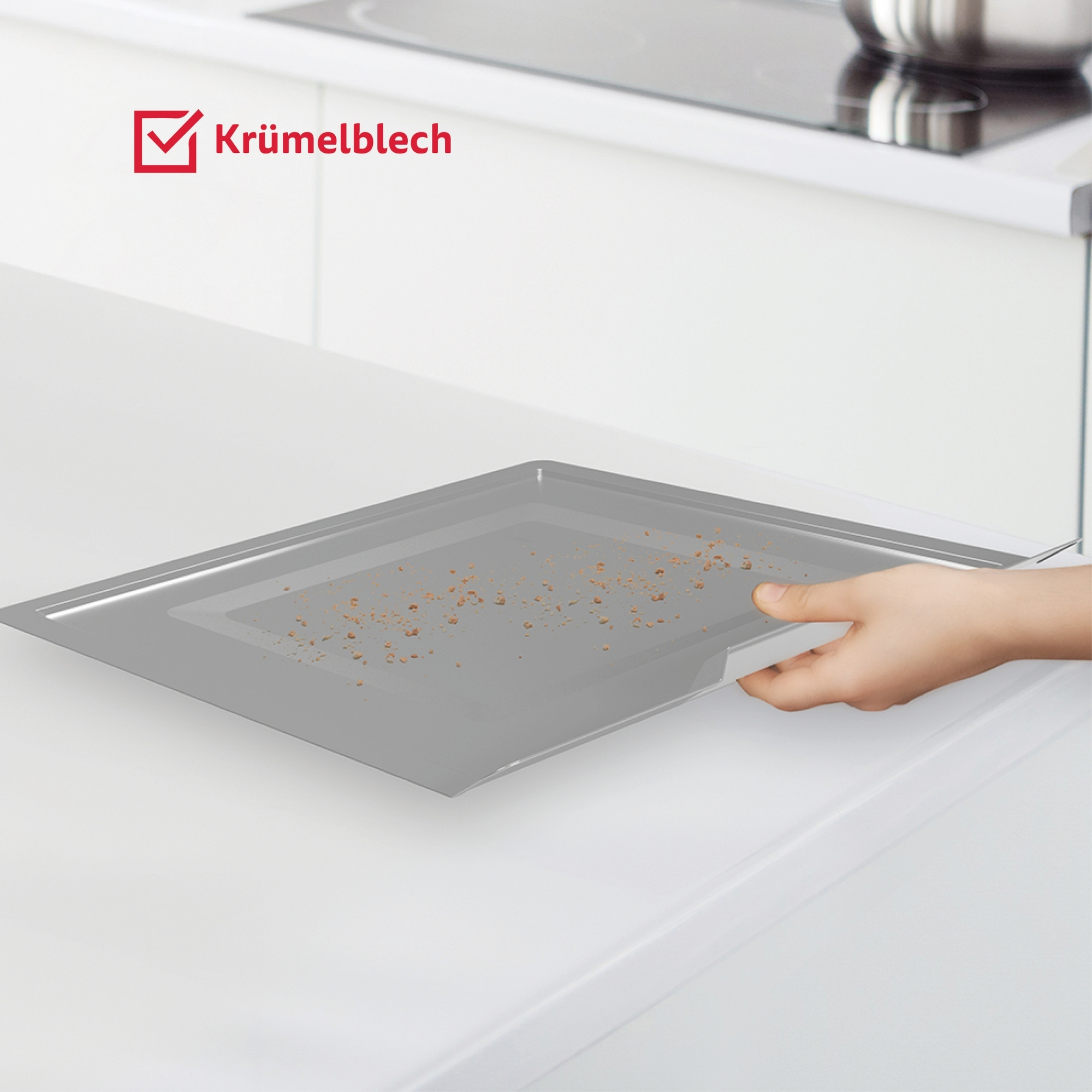 Una mano tiene un vassoio raccogli-briciole sopra un piano di lavoro bianco, con la scritta 'Krümelblech'.