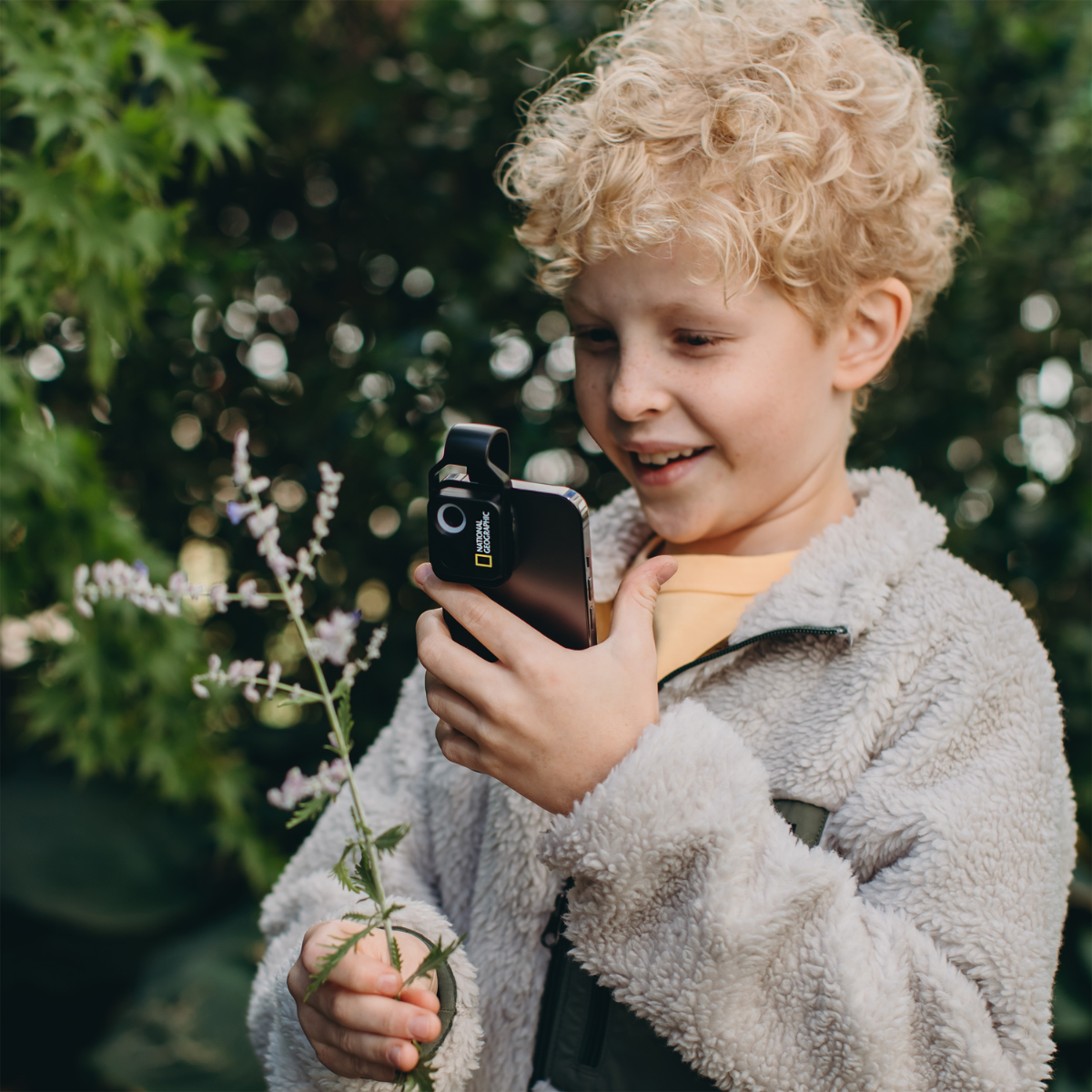 Niño sosteniendo un teléfono con un accesorio de cámara, apuntando a una planta. El niño sonríe.