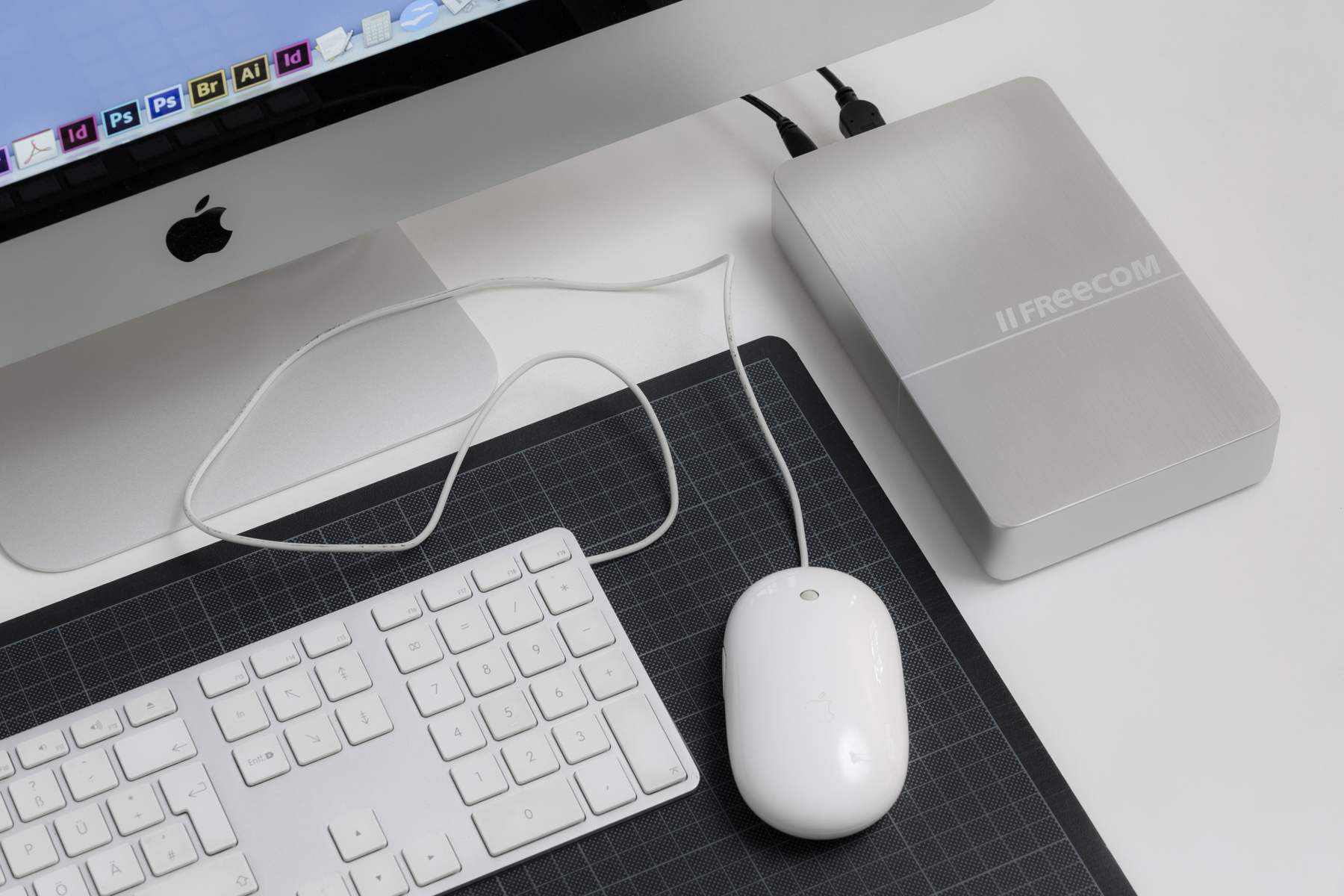 Konfiguracja komputera z klawiaturą, myszą i zewnętrznym dyskiem twardym. Elementy na czarnej macie, białym biurku.