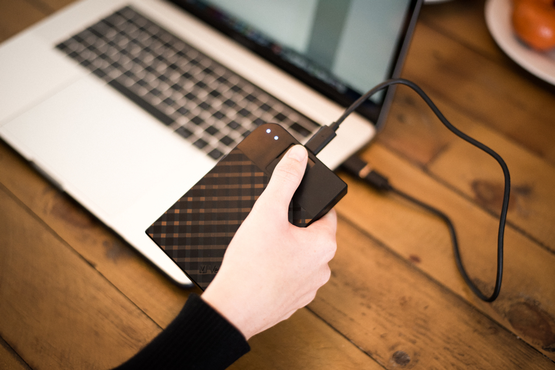 Ręka osoby podłącza czarny zewnętrzny dysk twardy do laptopa. Tło to drewniana powierzchnia.