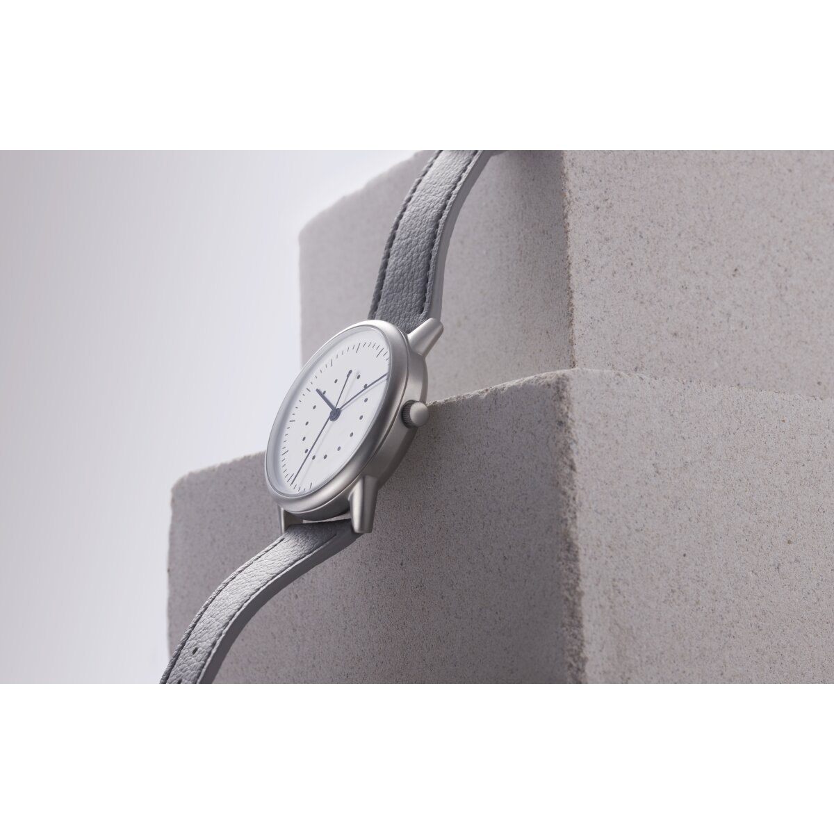 Silberne Uhr mit grauem Lederarmband. Die Uhr liegt auf grauen Steinblöcken, schräge Ansicht.