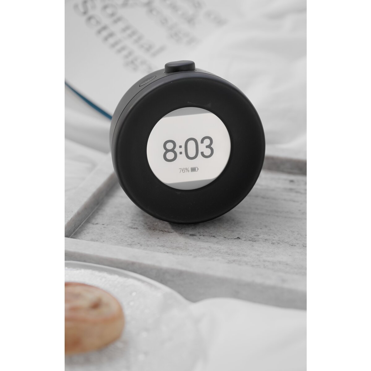 Zwarte klok toont 8:03 en 76% batterij. Het ligt op een grijze ondergrond, met een vage achtergrond.