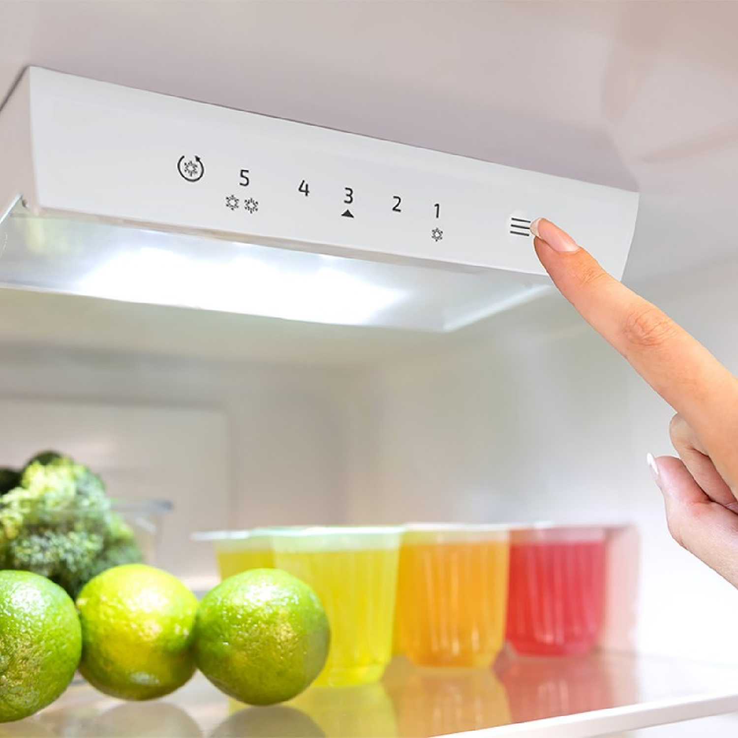Una mano tocando la configuración de un refrigerador. El fondo tiene frutas y jugos.
