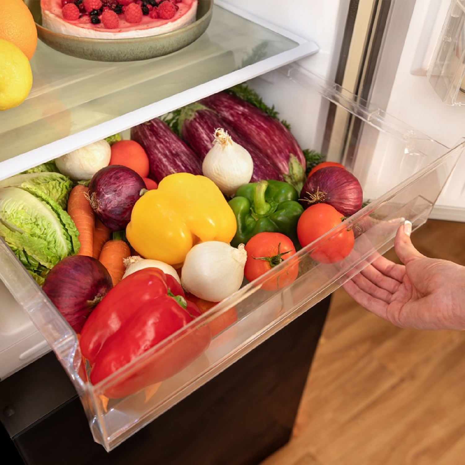 Un cajón de nevera lleno de verduras coloridas: pimientos, cebollas, zanahorias y tomates.