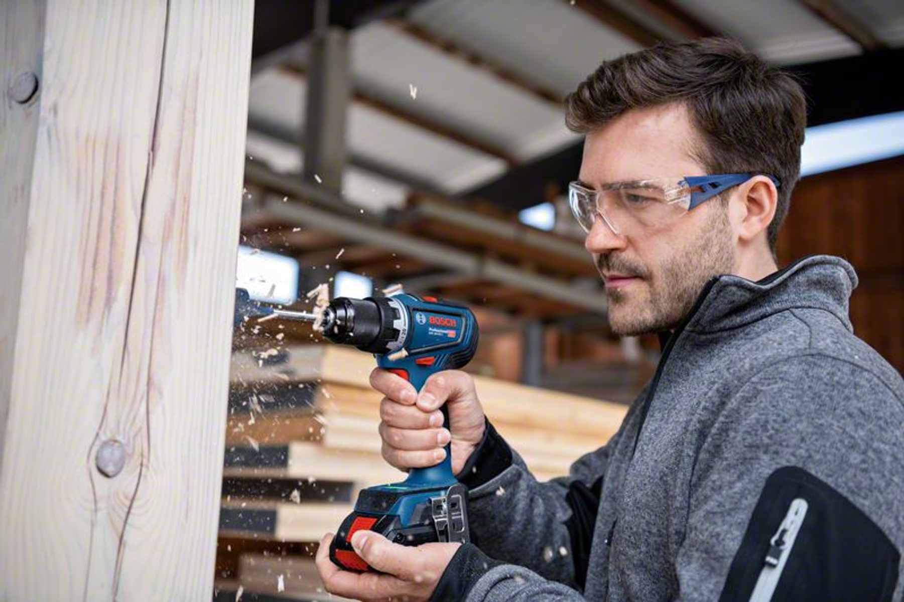 Un hombre con gafas de seguridad taladra una viga de madera con un taladro Bosch.