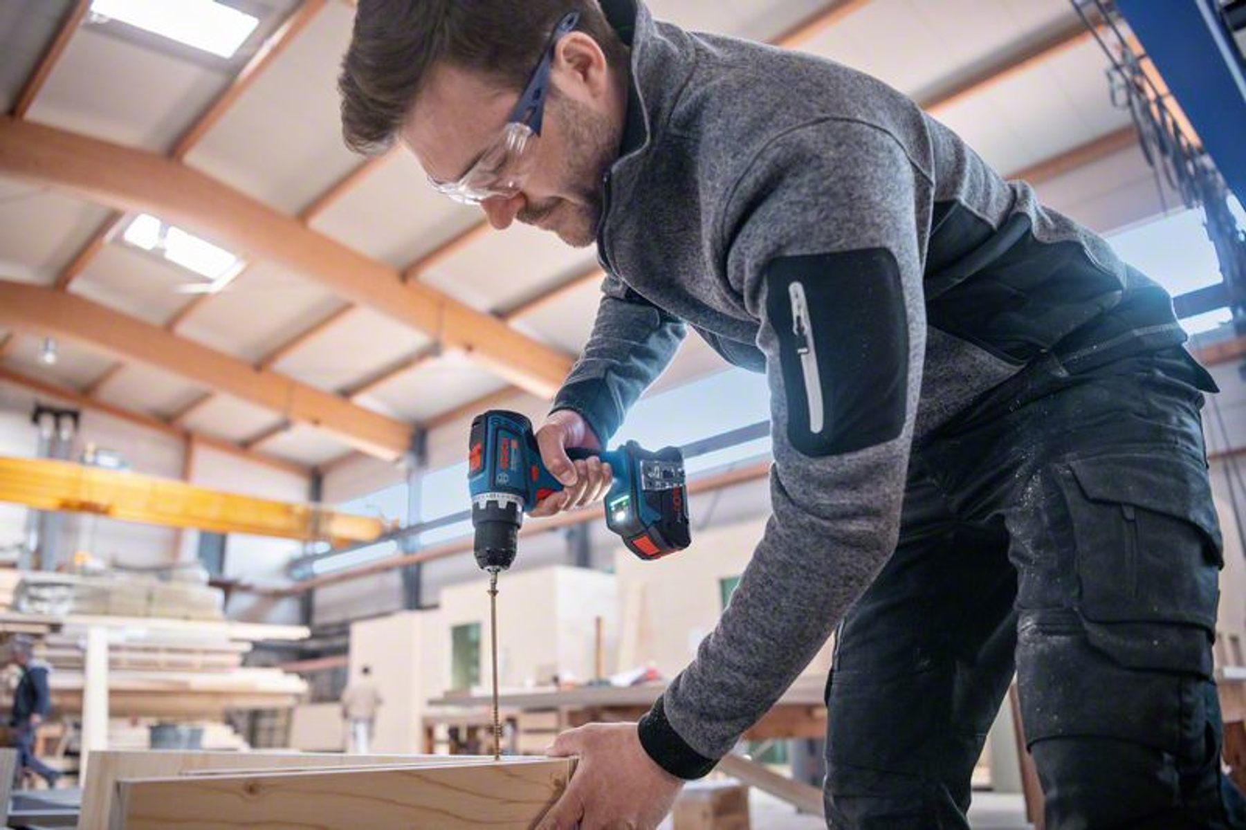 Un hombre taladra madera con un taladro eléctrico, usando gafas de seguridad. Taller.