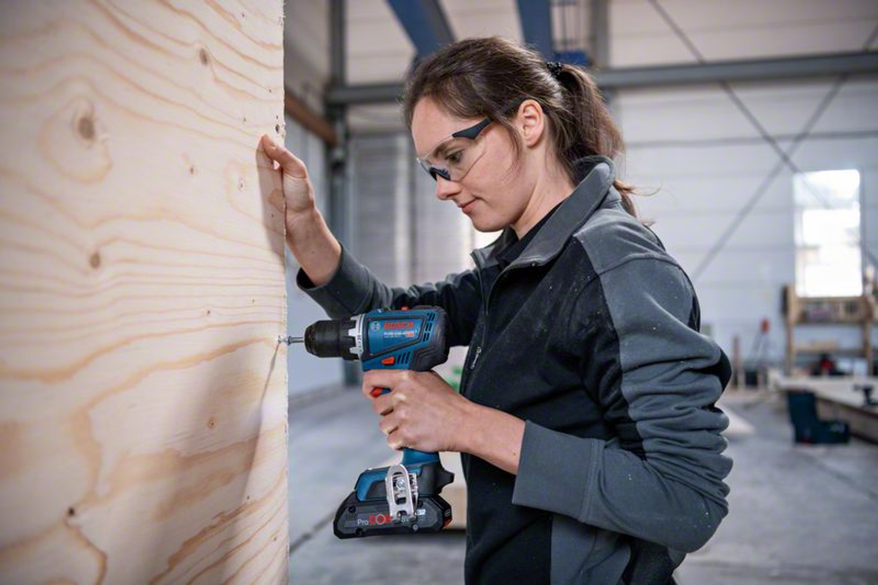 Mujer usando un taladro en madera, con gafas de seguridad, con un edificio al fondo.