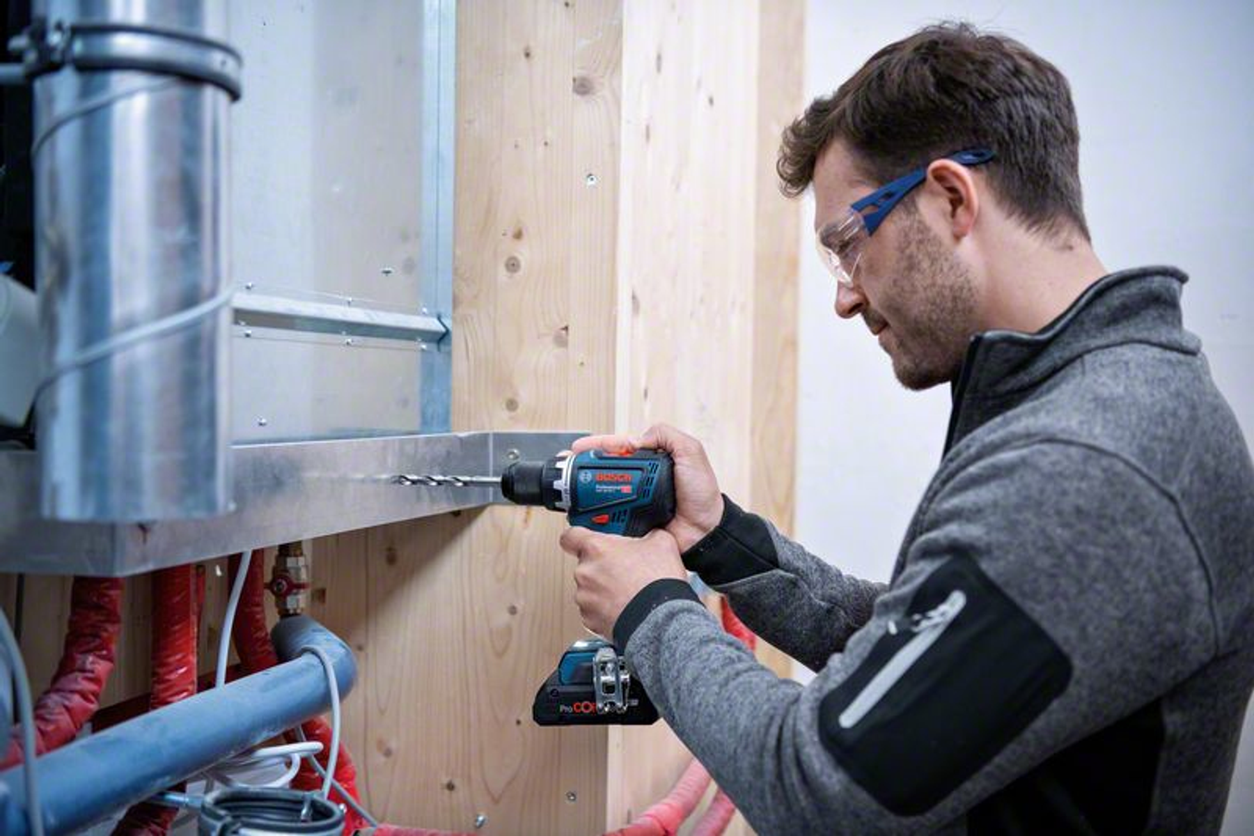 Hombre con gafas de seguridad taladrando un objeto metálico, trabajando en interiores.