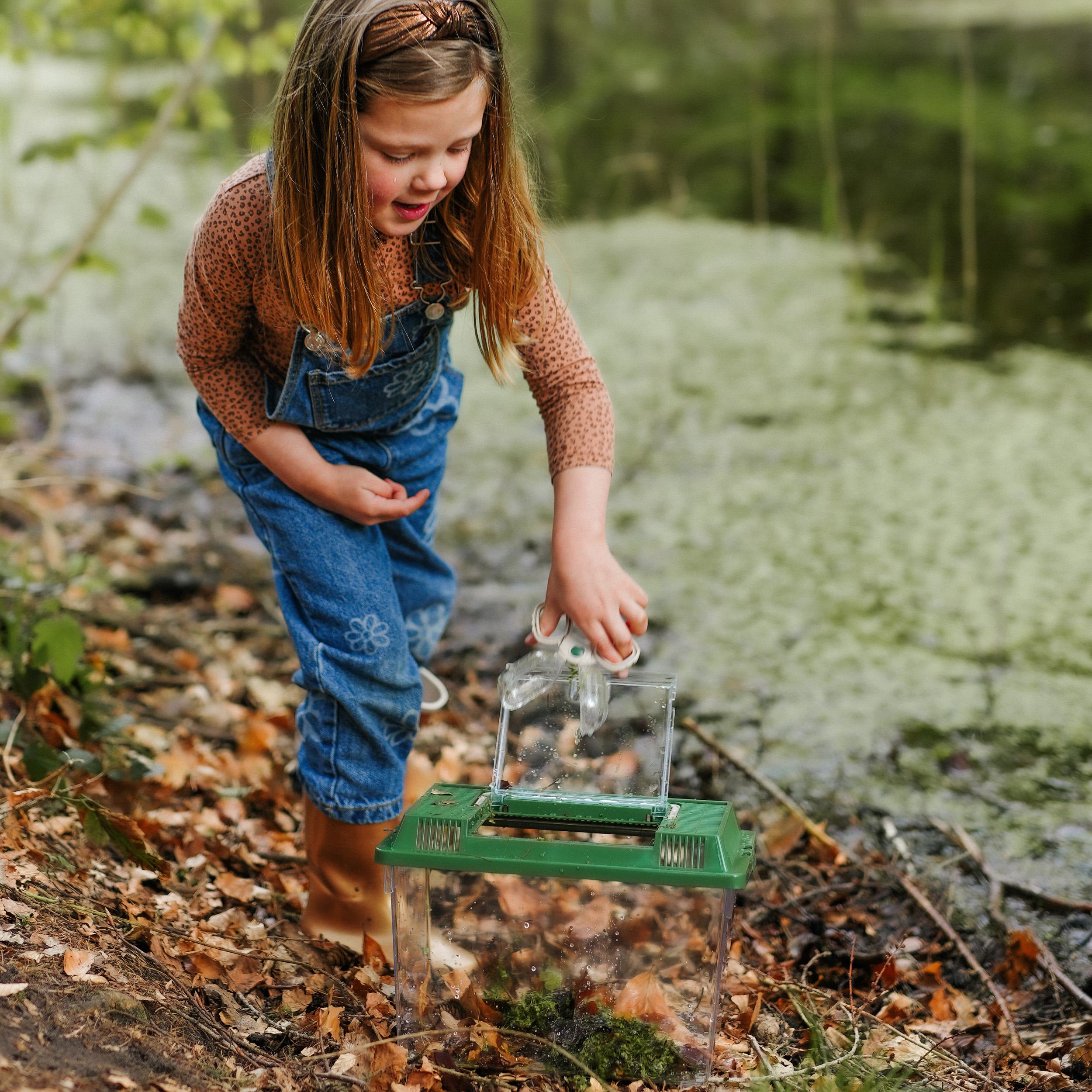 Una niña alcanza un recipiente verde en un estanque, mirando su contenido. Hojas marrones.