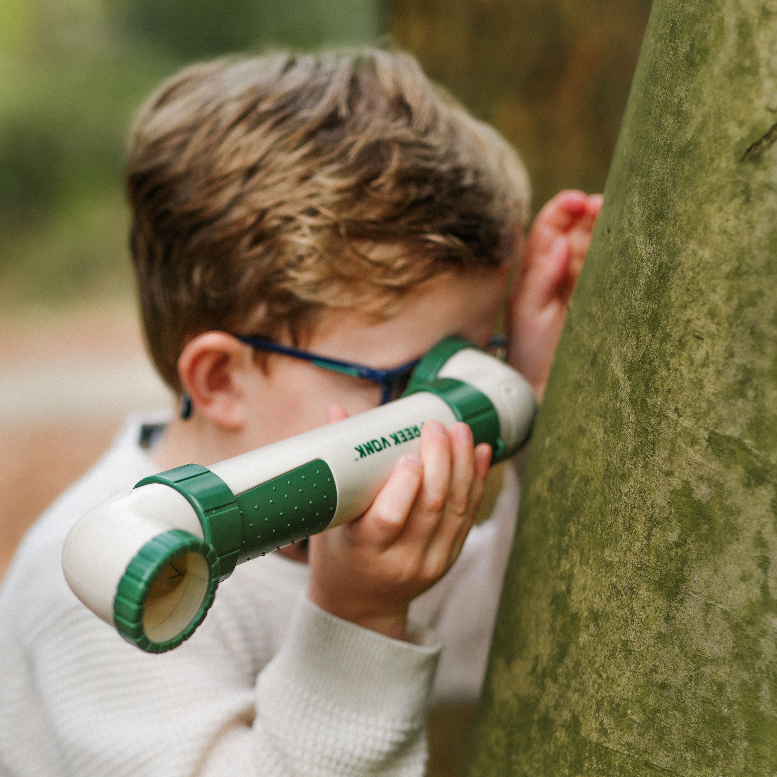Un niño mirando a través de un visor verde y blanco, cerca de un árbol. El niño lleva gafas.