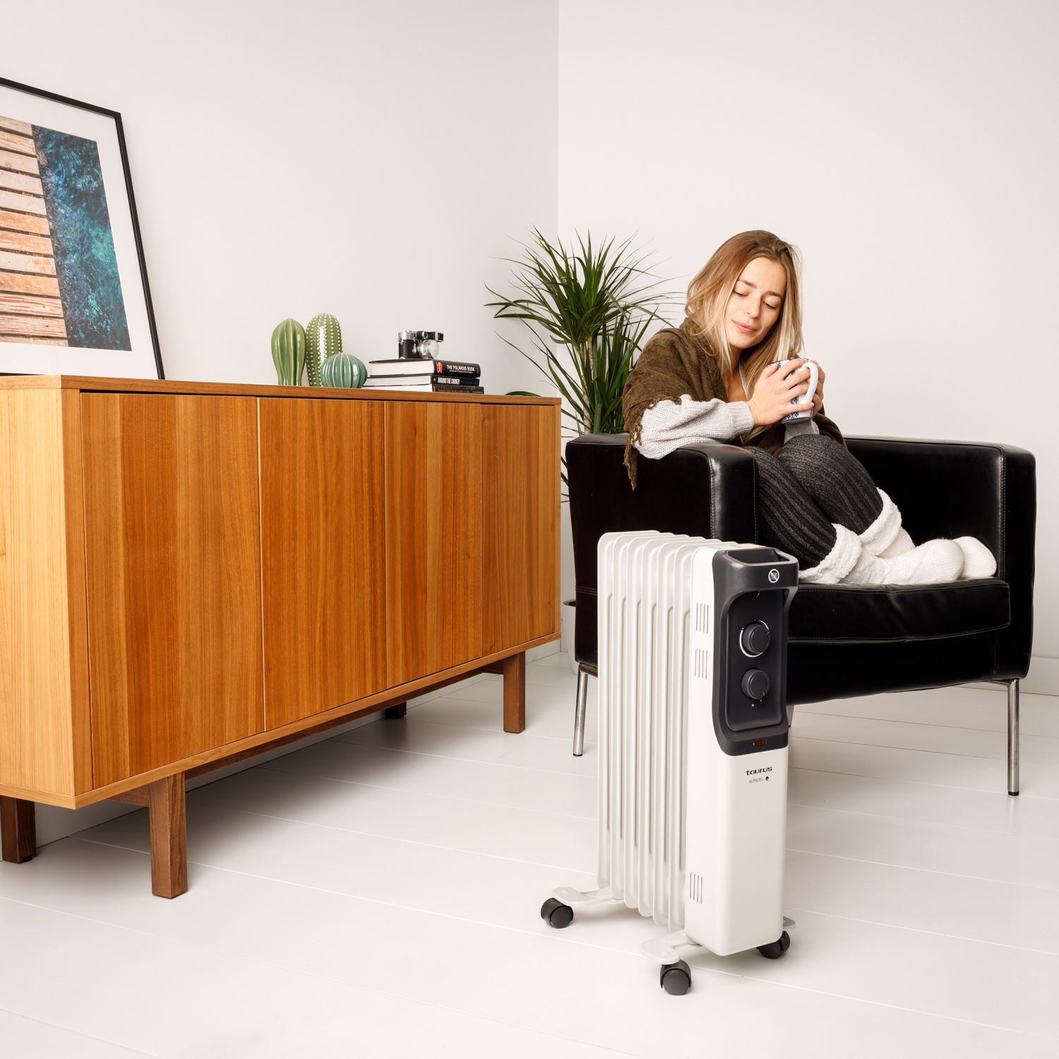Kobieta siedzi na czarnym krześle, trzymając kubek. Biały grzejnik stoi przed nią.
