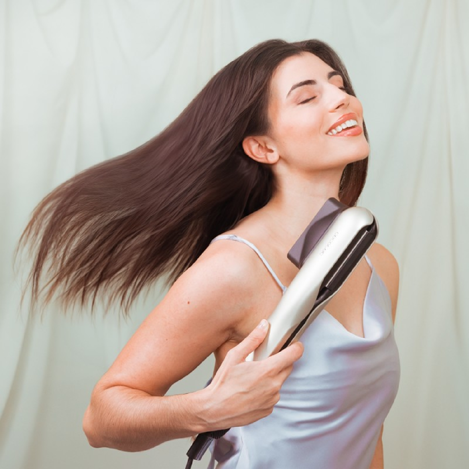 Mujer sonriendo, sosteniendo una plancha para el cabello con el pelo suelto, fondo claro, en una habitación.