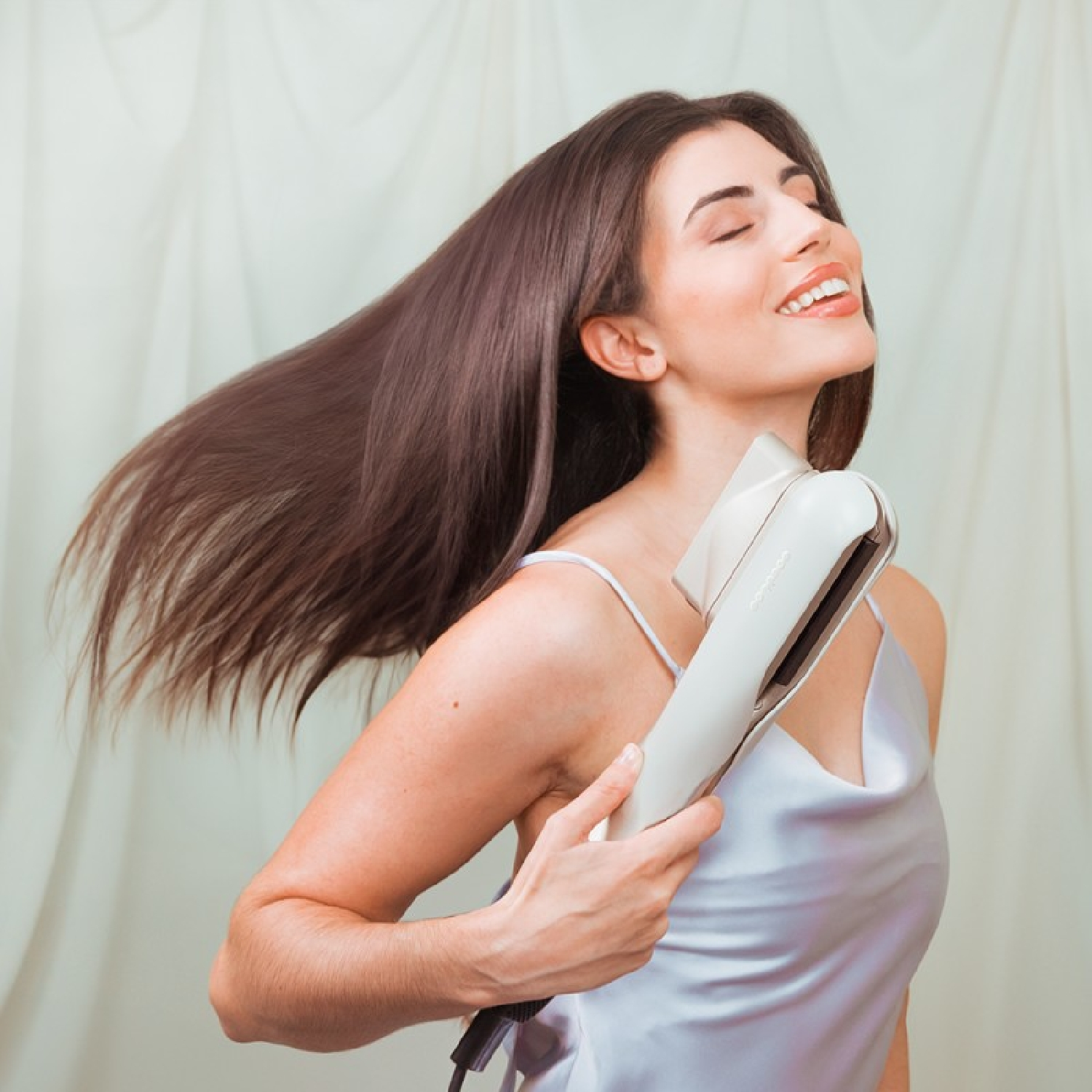 Frau mit langen braunen Haaren benutzt einen Haarglätter. Sie lächelt und ihre Haare fließen.
