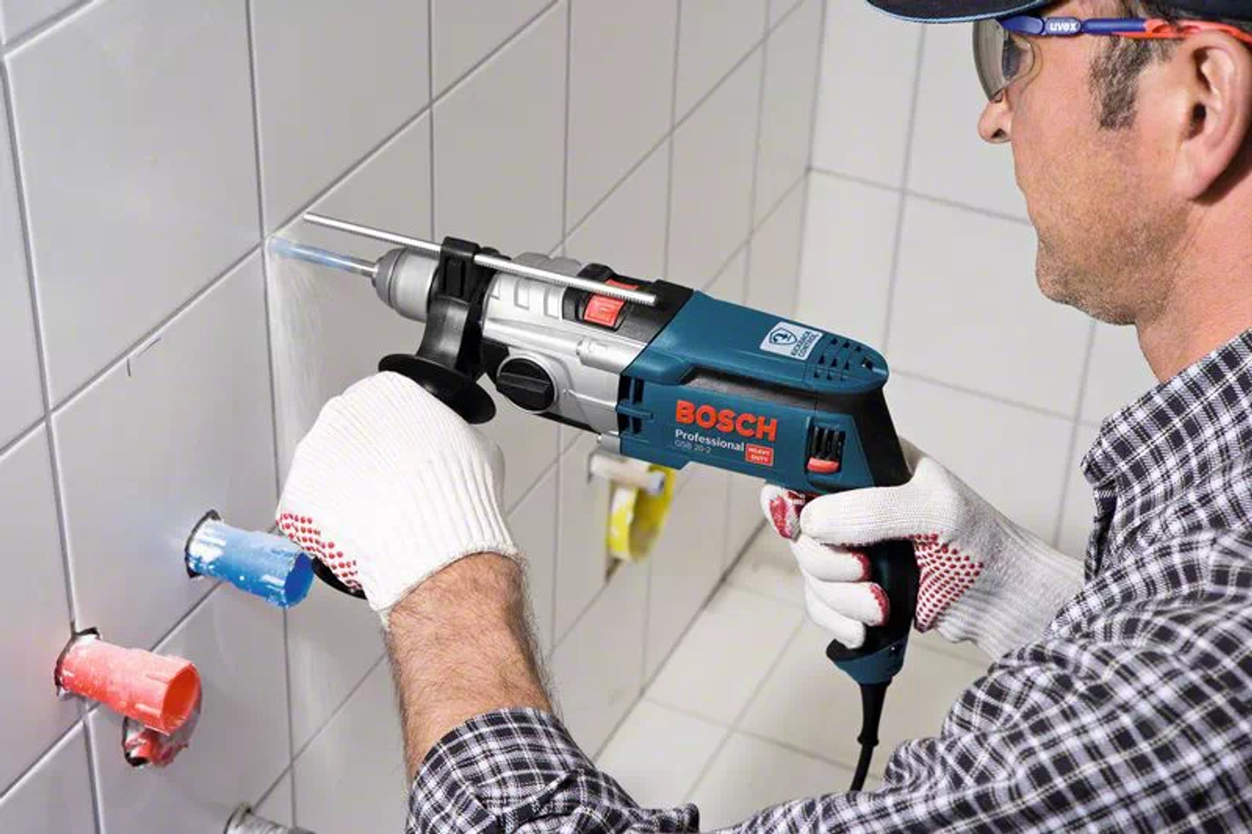 Mann bohrt mit Bosch-Bohrmaschine in geflieste Wand, trägt Schutzbrille und Handschuhe.