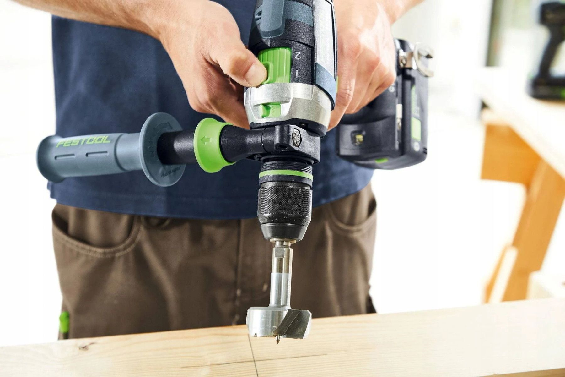 Osoba używająca wiertarki na drewnianej powierzchni. Wiertarka jest czarna i zielona. Ręka trzyma wiertarkę.