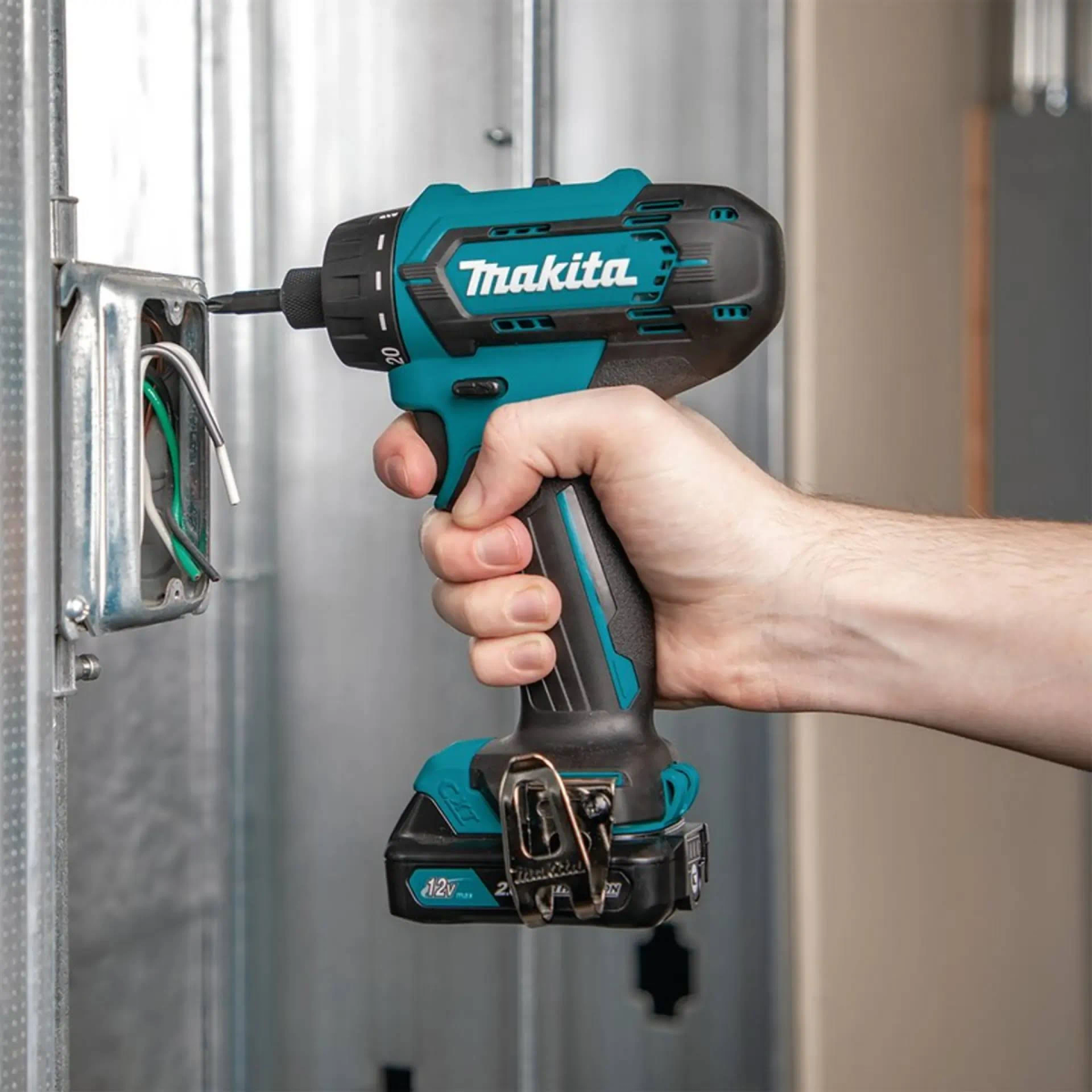 Mano sosteniendo un taladro Makita azul y negro, taladrando en una caja eléctrica. Fondo de metal.