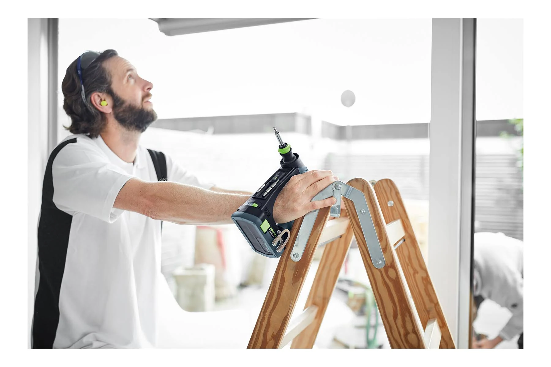 Mężczyzna na drabinie używający wiertarki. Patrzy w górę i nosi ochronę słuchu.